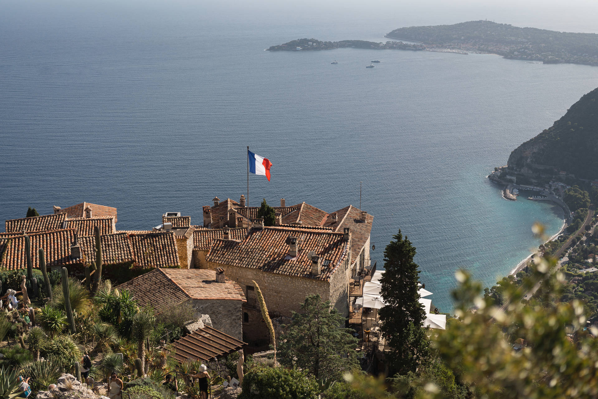 A French flag flies over a village with terracotta roofs in the foreground, overlooking a sparkling blue bay and distant coastline in the background.