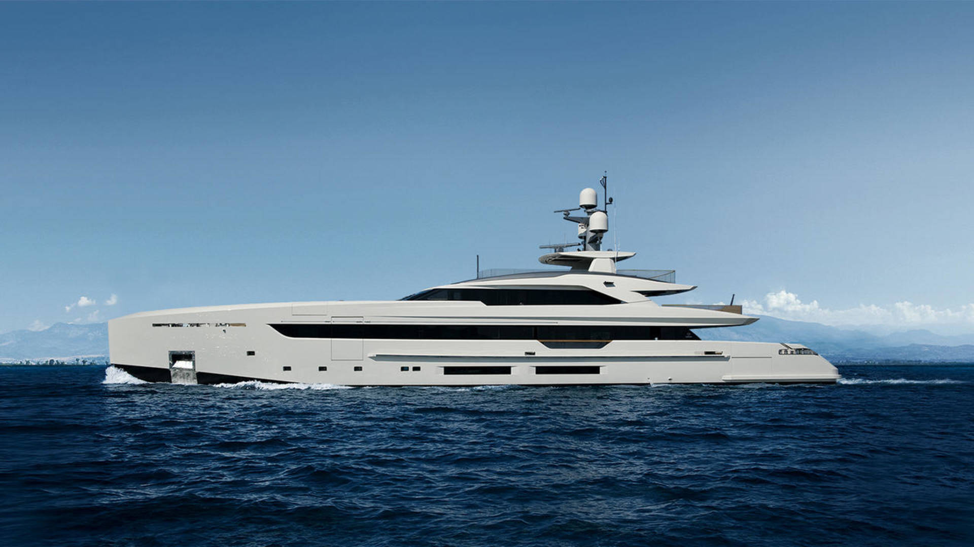 A sleek, modern superyacht glides through deep blue water with a hazy coastline and clear sky in the background.