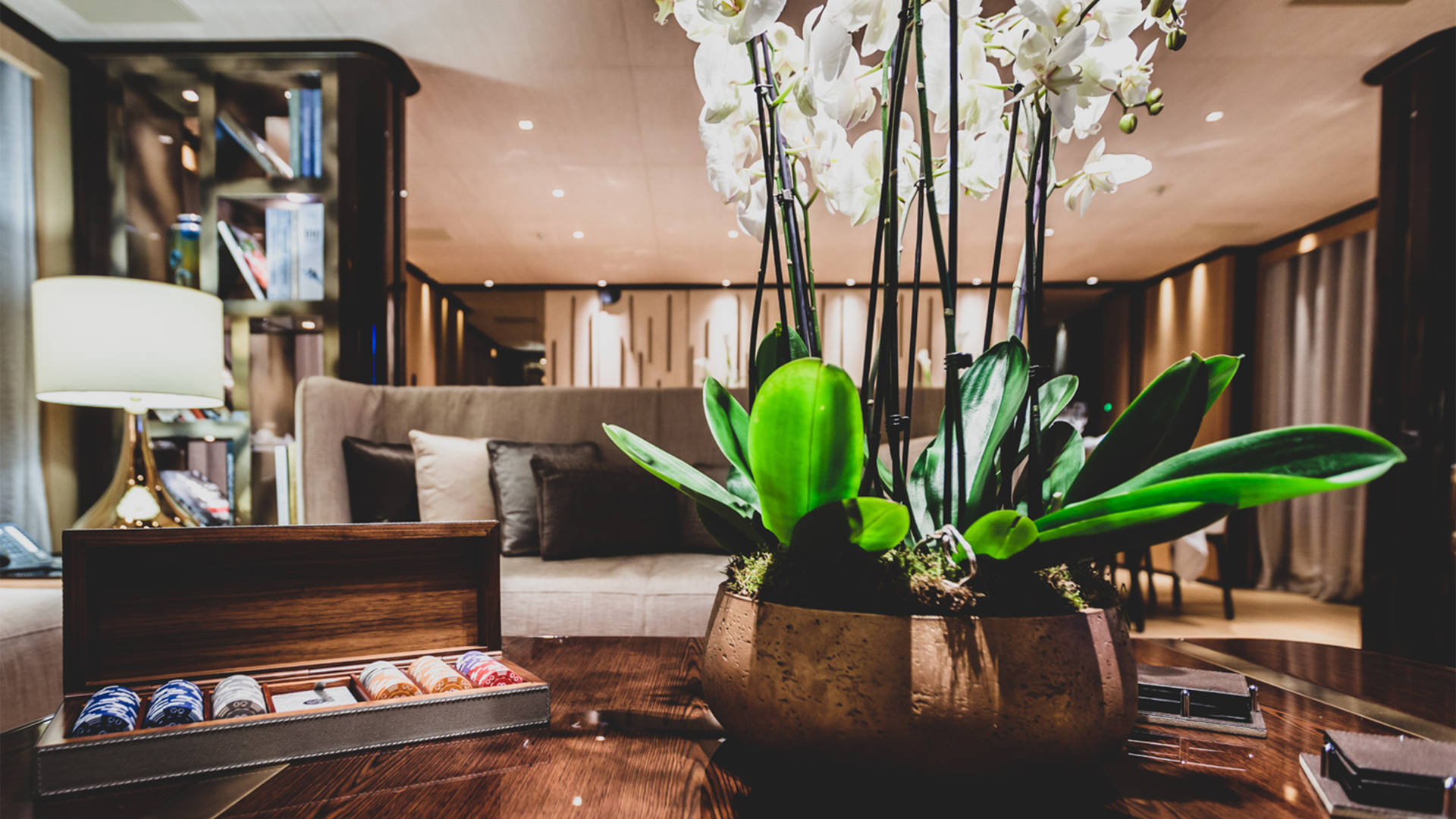 A potted orchid and a wooden box of poker chips rest on a polished table in the foreground, with a plush sofa and bookshelves visible in the softly lit background.