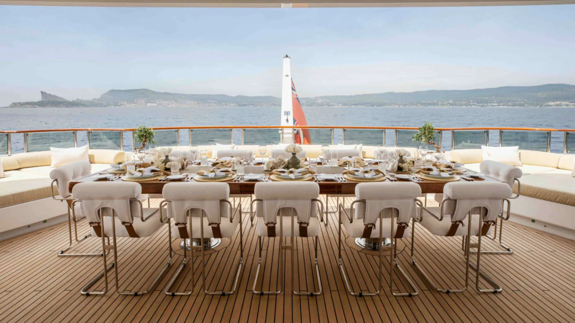 A long dining table set for a meal on a yacht's deck is surrounded by white chairs, with a calm sea and distant coastline visible in the background.