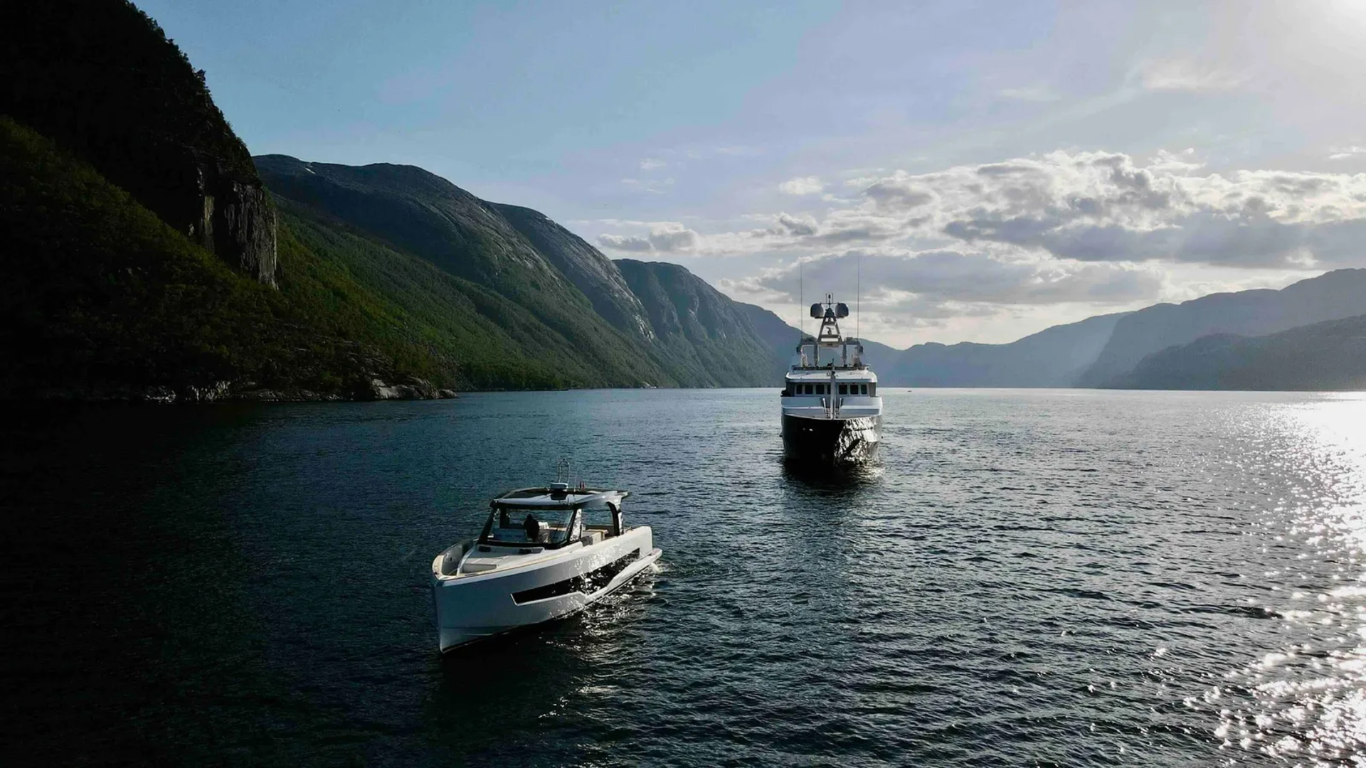 A sleek white speedboat cruises in the foreground of a calm, wide body of water, with a larger yacht following behind, all set against a backdrop of majestic, tree-covered mountains under a bright, cloudy sky.