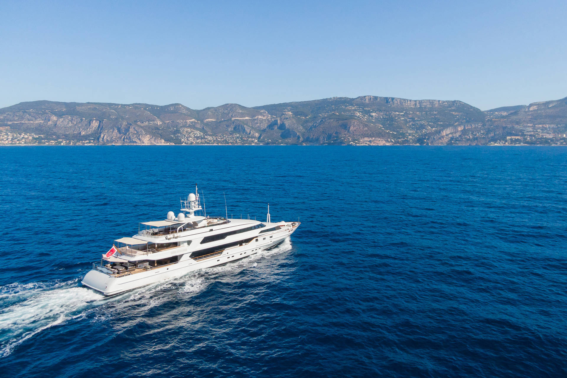 A large white yacht cruises through deep blue water with a mountainous coastline in the background.