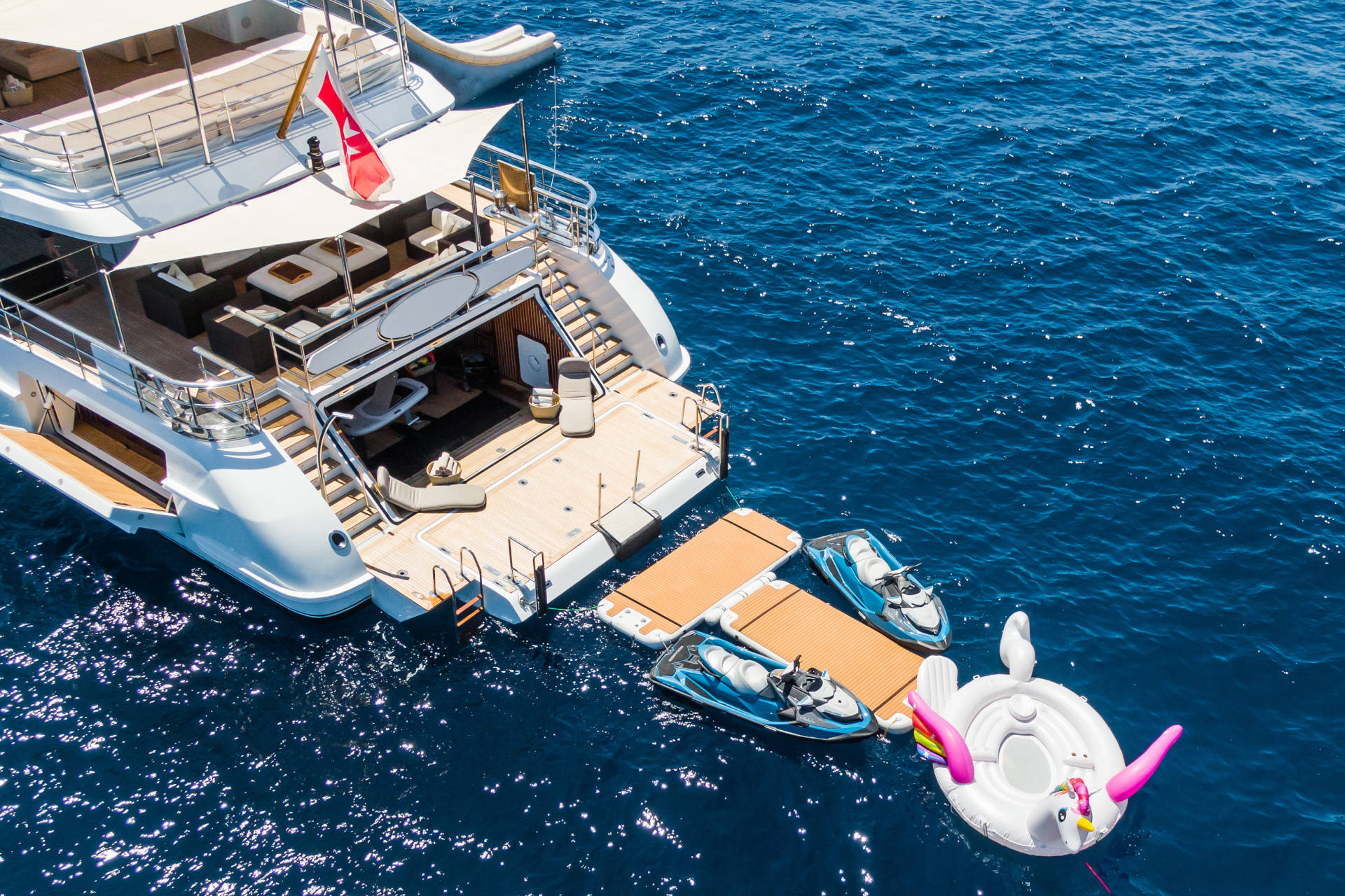 Two jet skis are docked at the rear of a luxurious yacht, with a pink flamingo float bobbing in the deep blue ocean.