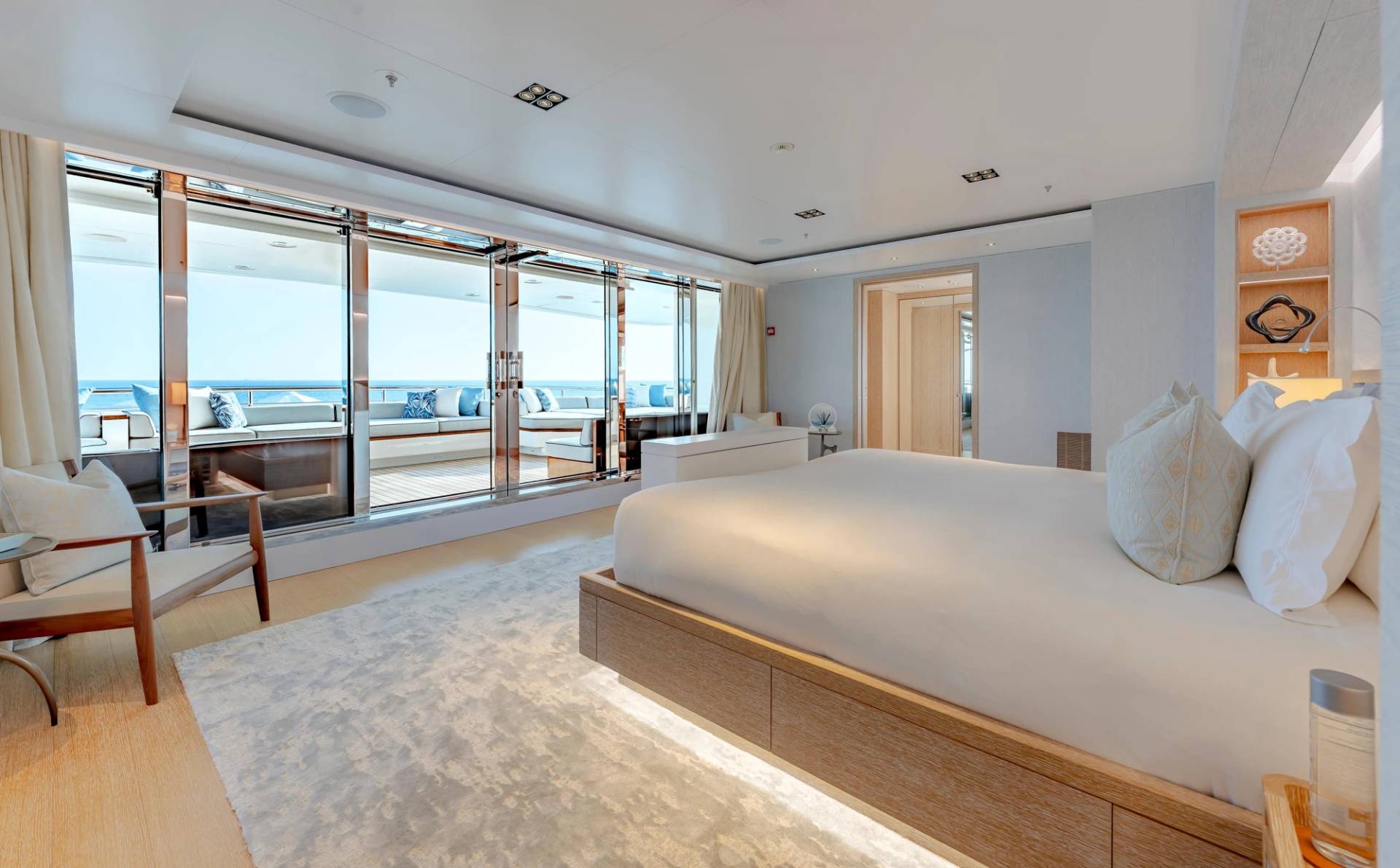 A luxurious bedroom with a large bed in the foreground opens onto a sunlit balcony with ocean views in the background.