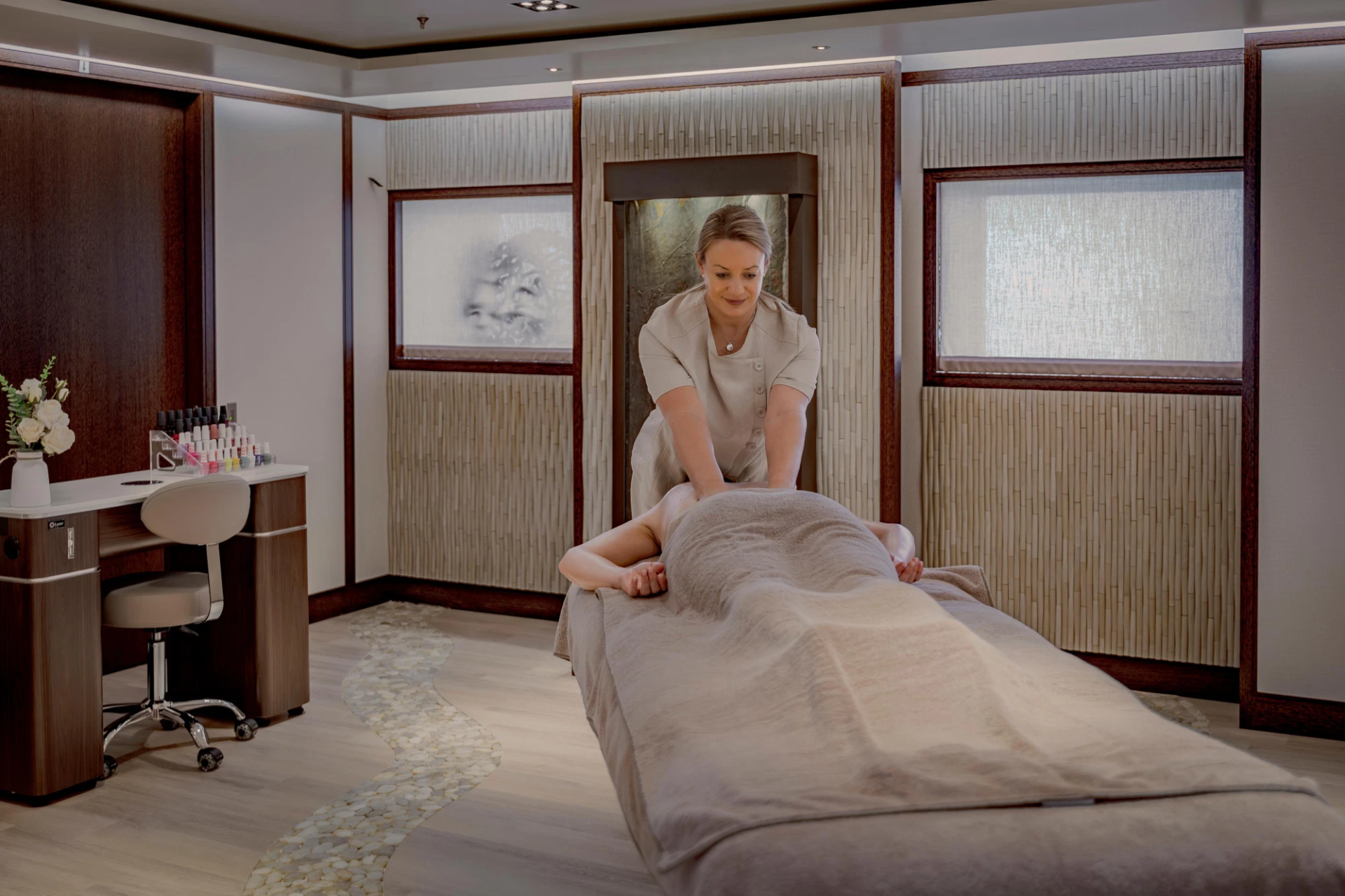 A massage therapist works on a client lying on a massage table in a spa room with a nail station in the foreground and textured walls with artwork in the background.