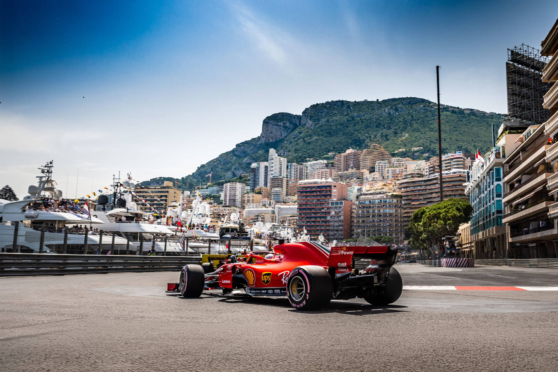 A red Formula 1 car turns a corner on a sunny street circuit with yachts and buildings in the background.