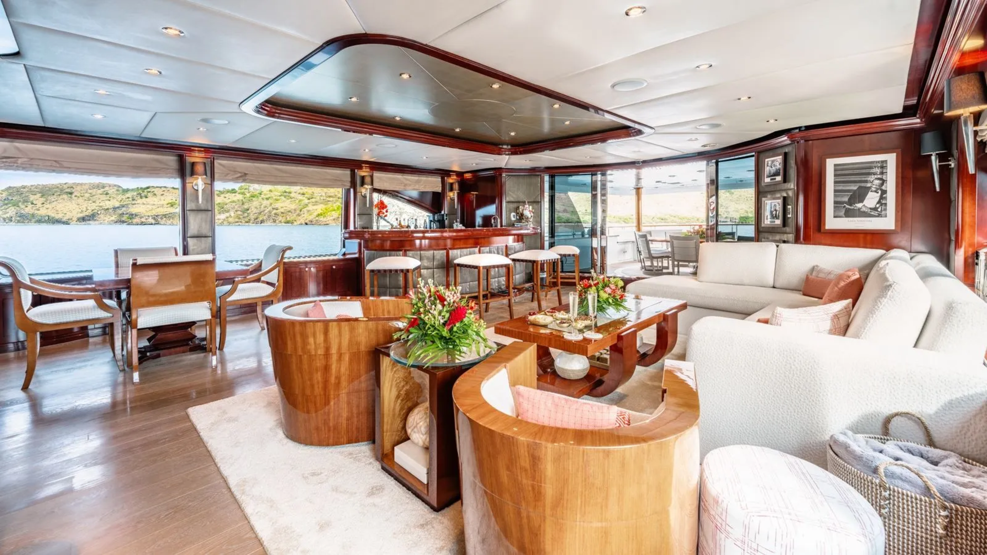 Luxurious yacht interior with plush seating and a bar in the foreground, overlooking a serene body of water and lush green hills in the background.