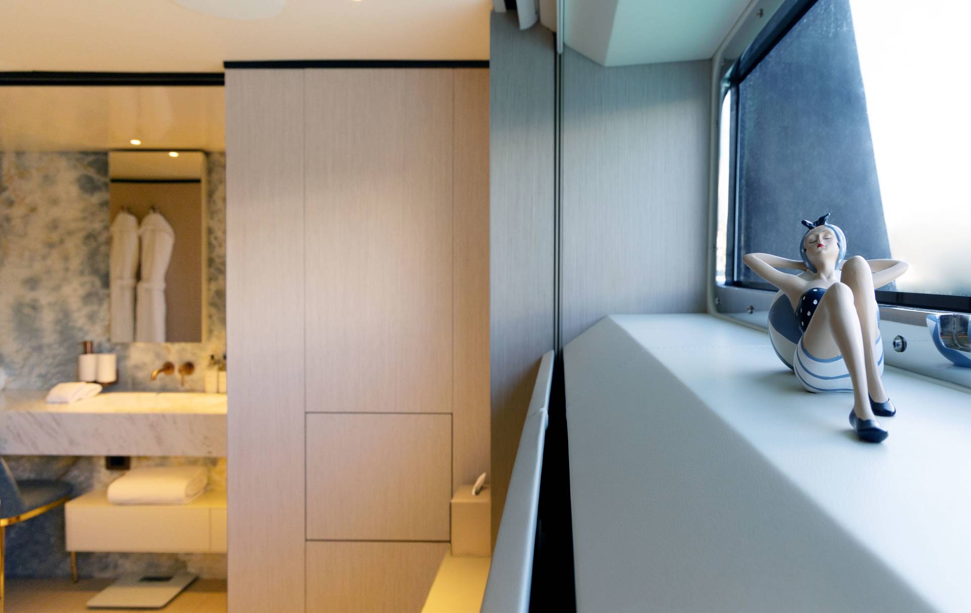 A miniature figure relaxes by a large window in the foreground, while a modern bathroom with marble accents is visible in the background.