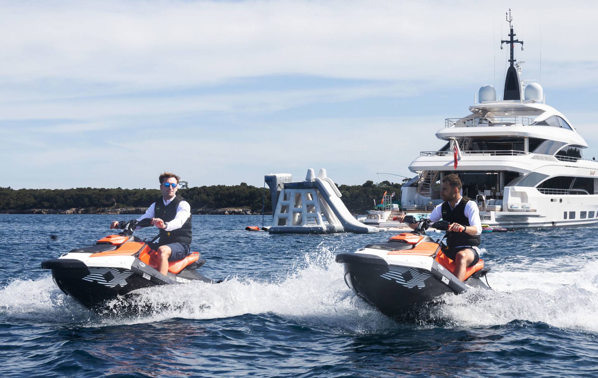 Two men ride jet skis in the foreground, leaving wakes in the water, while a large yacht and a floating water slide are visible in the background.