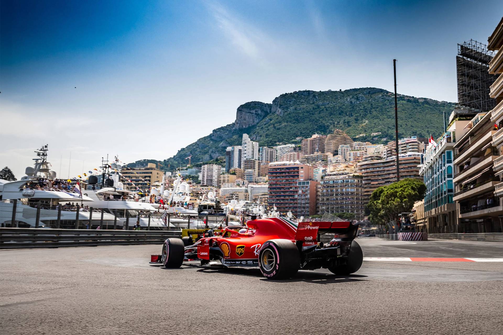 A red Formula 1 car races on a street circuit with yachts and a city skyline in the background.