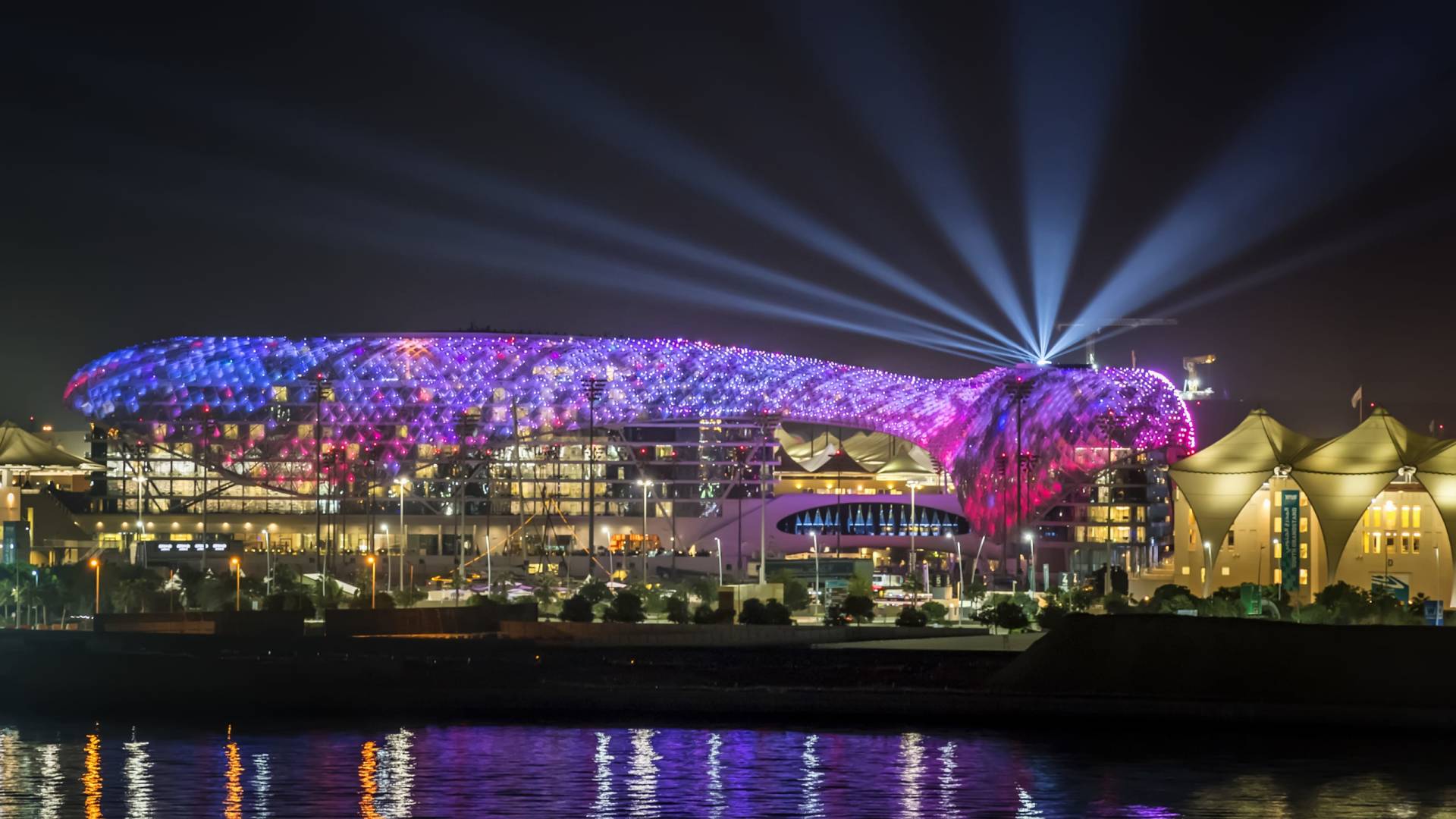 A brightly lit, colorful stadium with a distinctive curved roof stands illuminated against a dark night sky, with its reflection shimmering on the calm water in the foreground.
