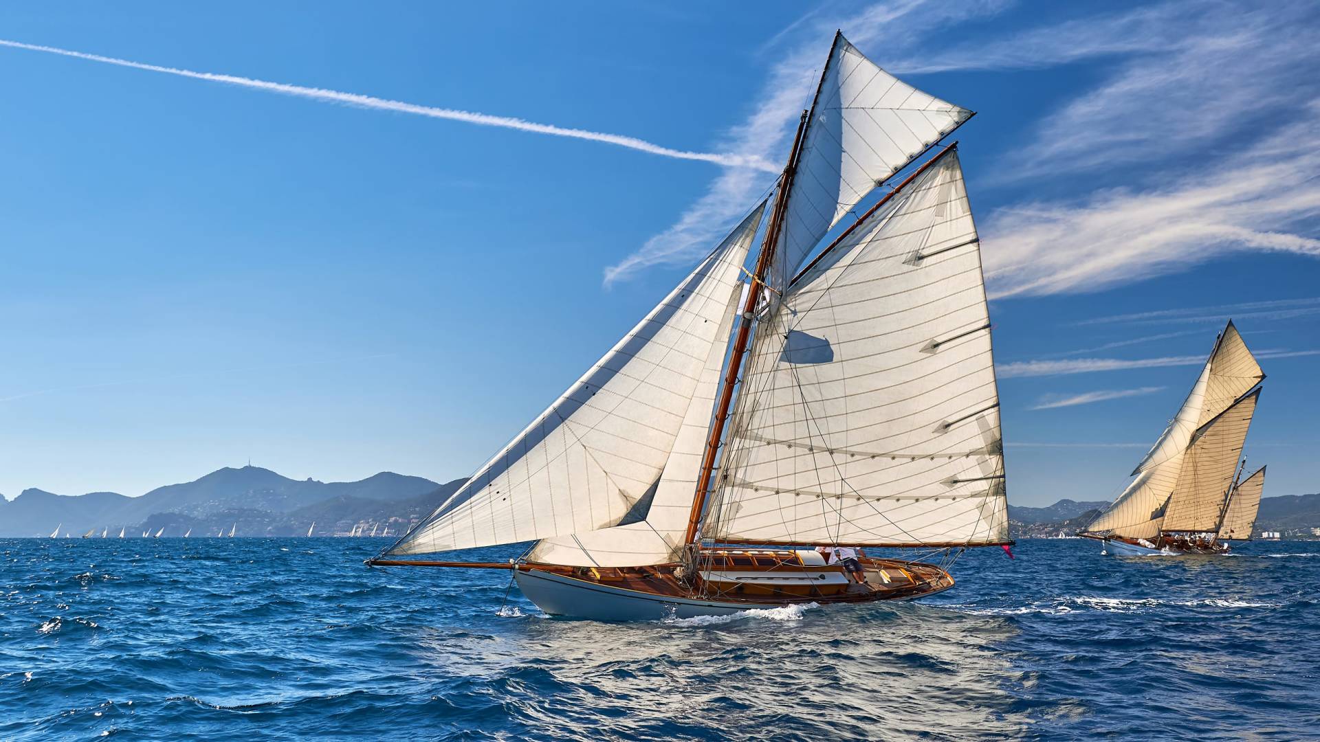 A classic sailboat with white sails glides across a blue ocean, with other sailboats and a distant coastline under a clear sky.