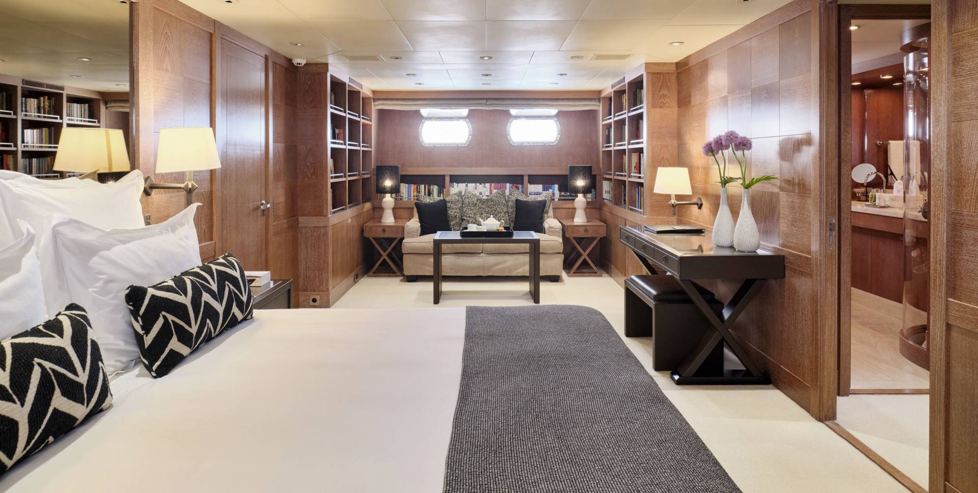 A luxurious bedroom with a large bed in the foreground and a desk and seating area in the background, all surrounded by wooden paneling and bookshelves.