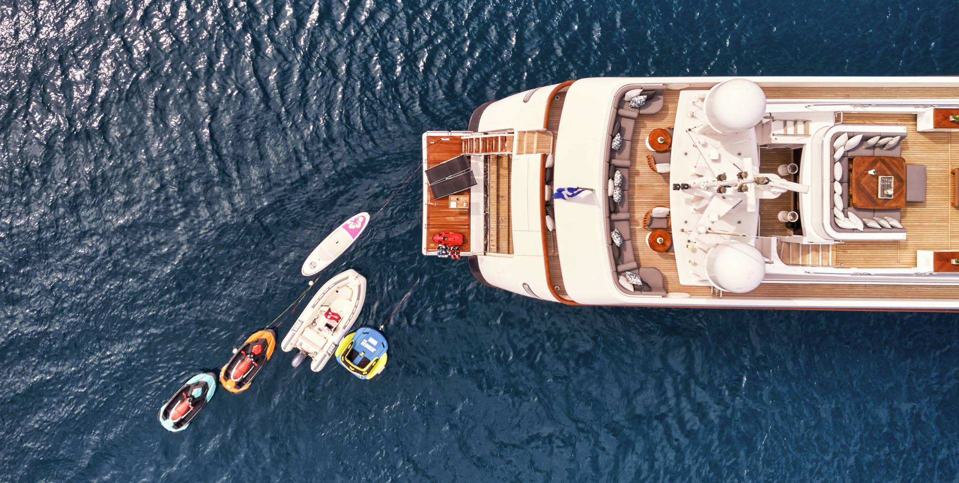 A large yacht floats on the deep blue sea, with several inflatable water toys trailing behind it.