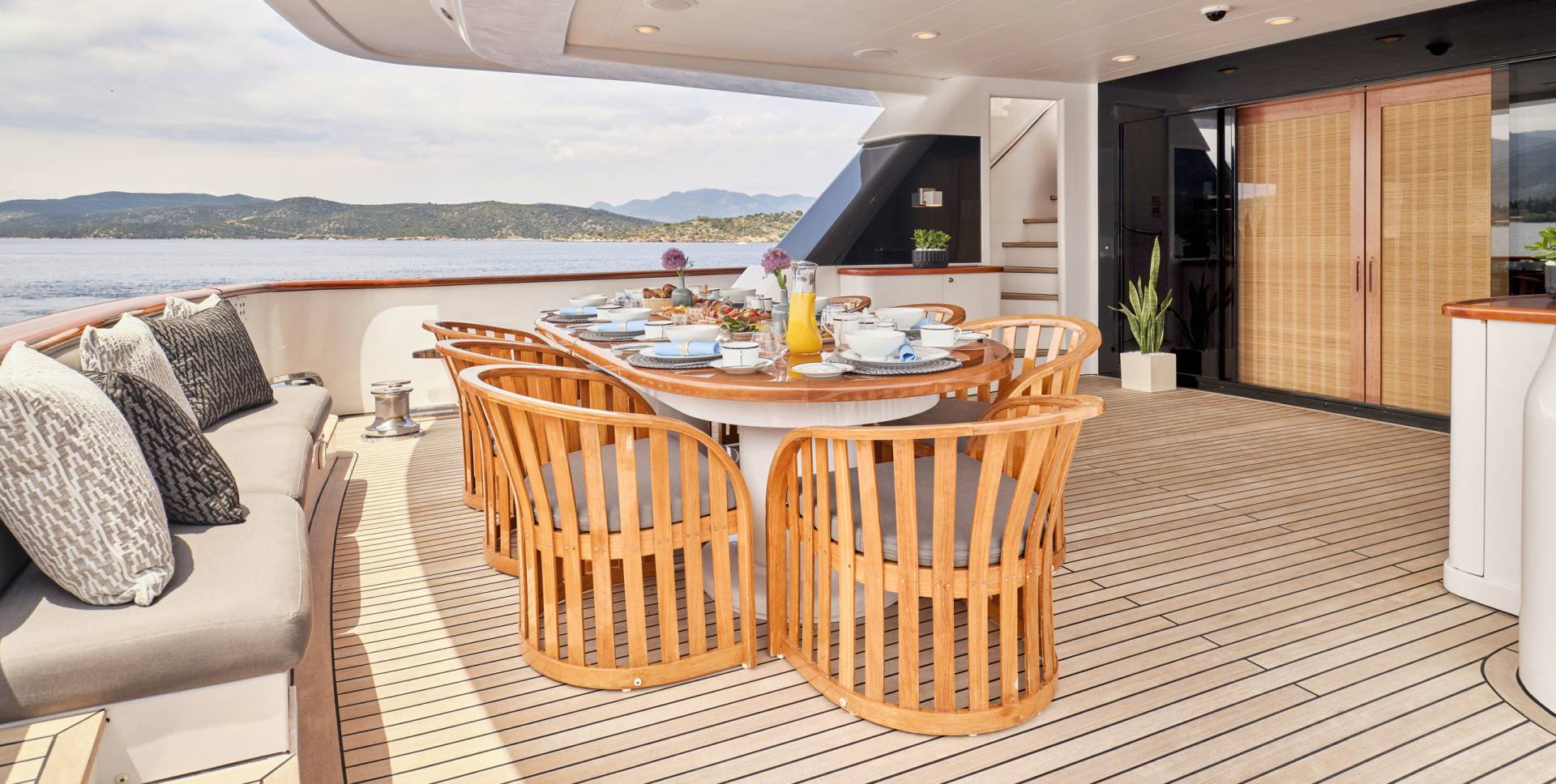 A beautifully set dining table with chairs on a yacht's deck overlooks a serene coastal landscape.