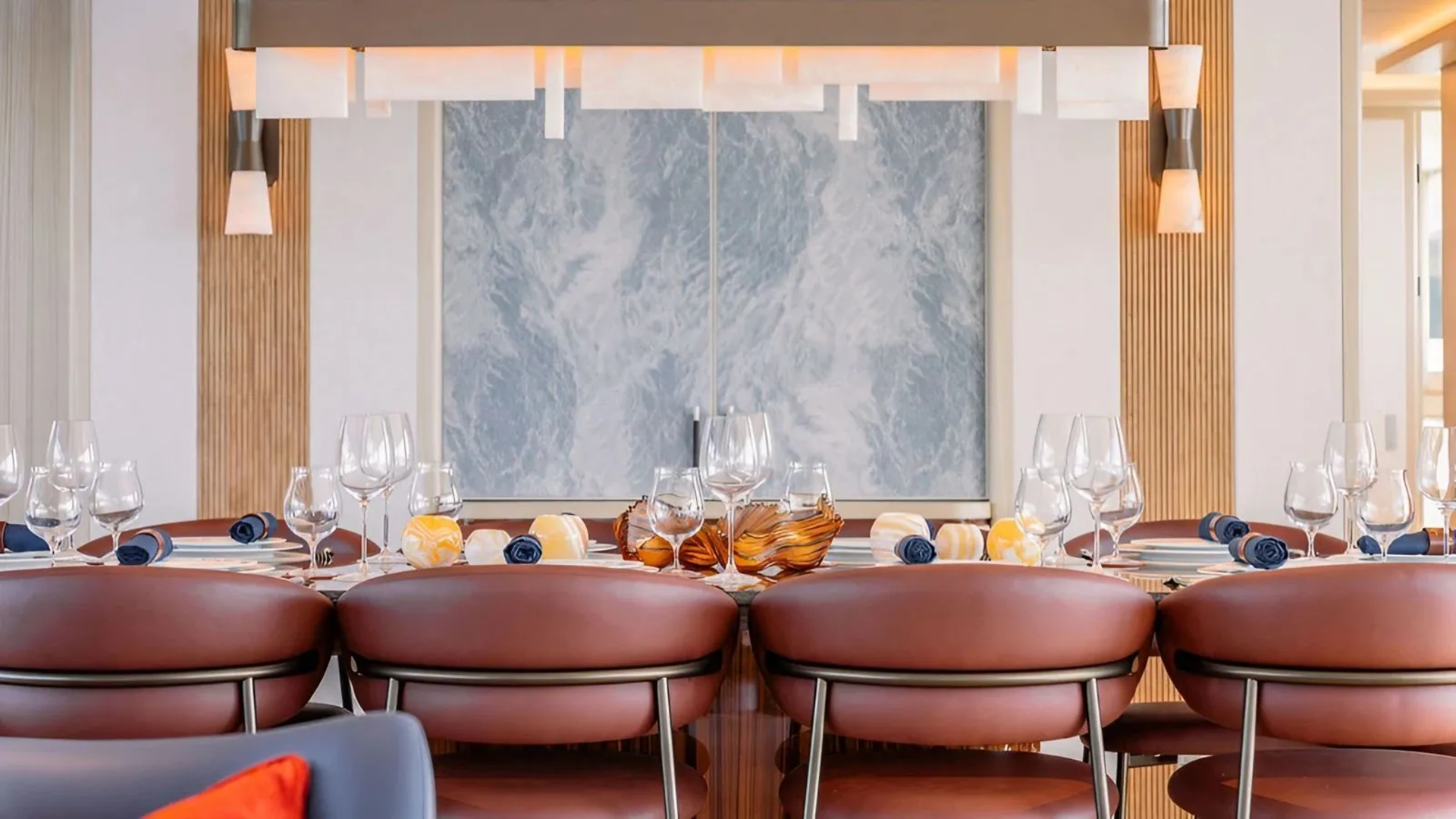 A formal dining table with brown leather chairs in the foreground is set with glassware and place settings, while a marbled wall and modern light fixture adorn the background.