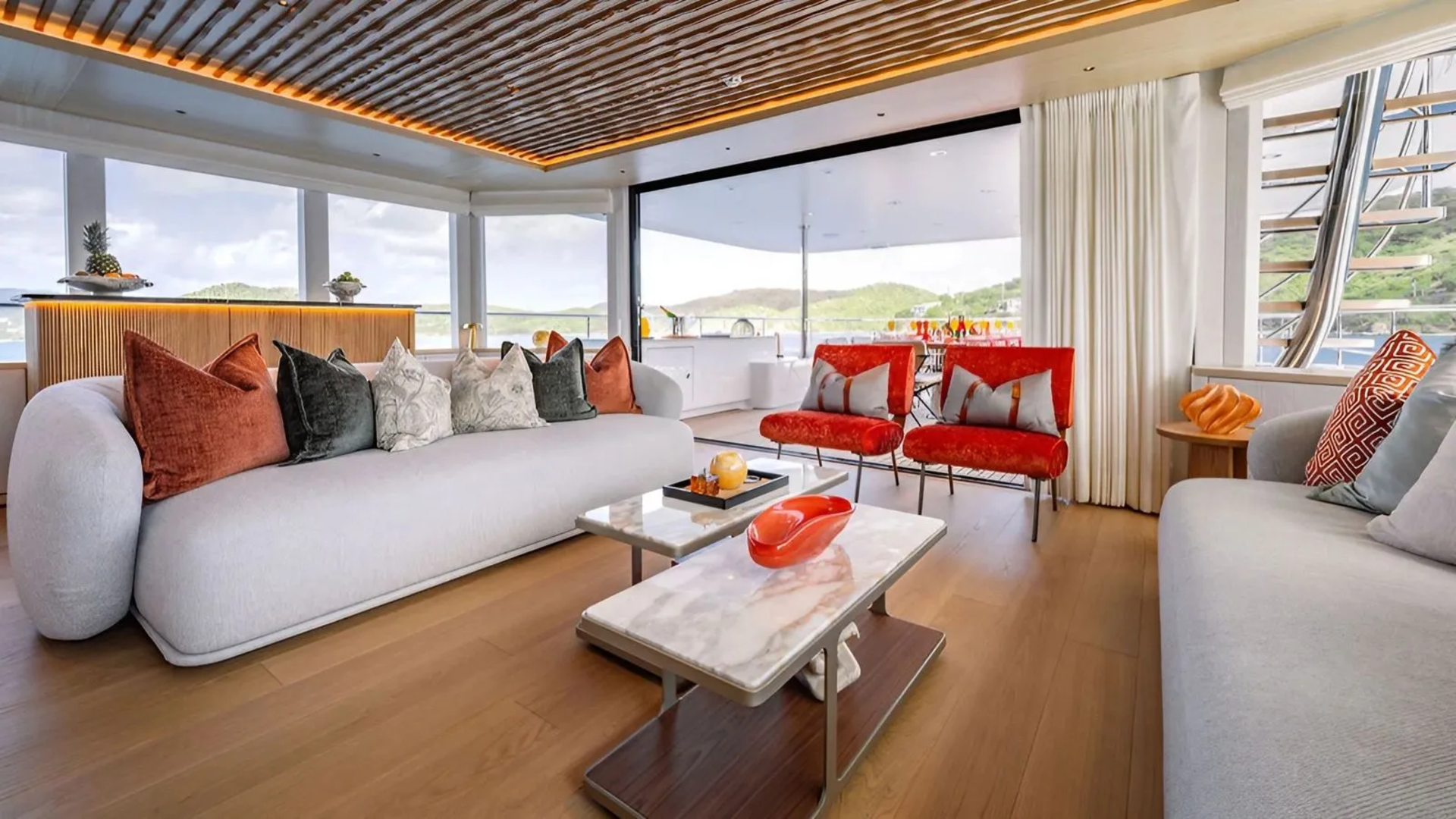 A luxurious yacht interior with plush white sofas and colorful accent chairs in the foreground, and a scenic view of rolling hills and water through large windows in the background.