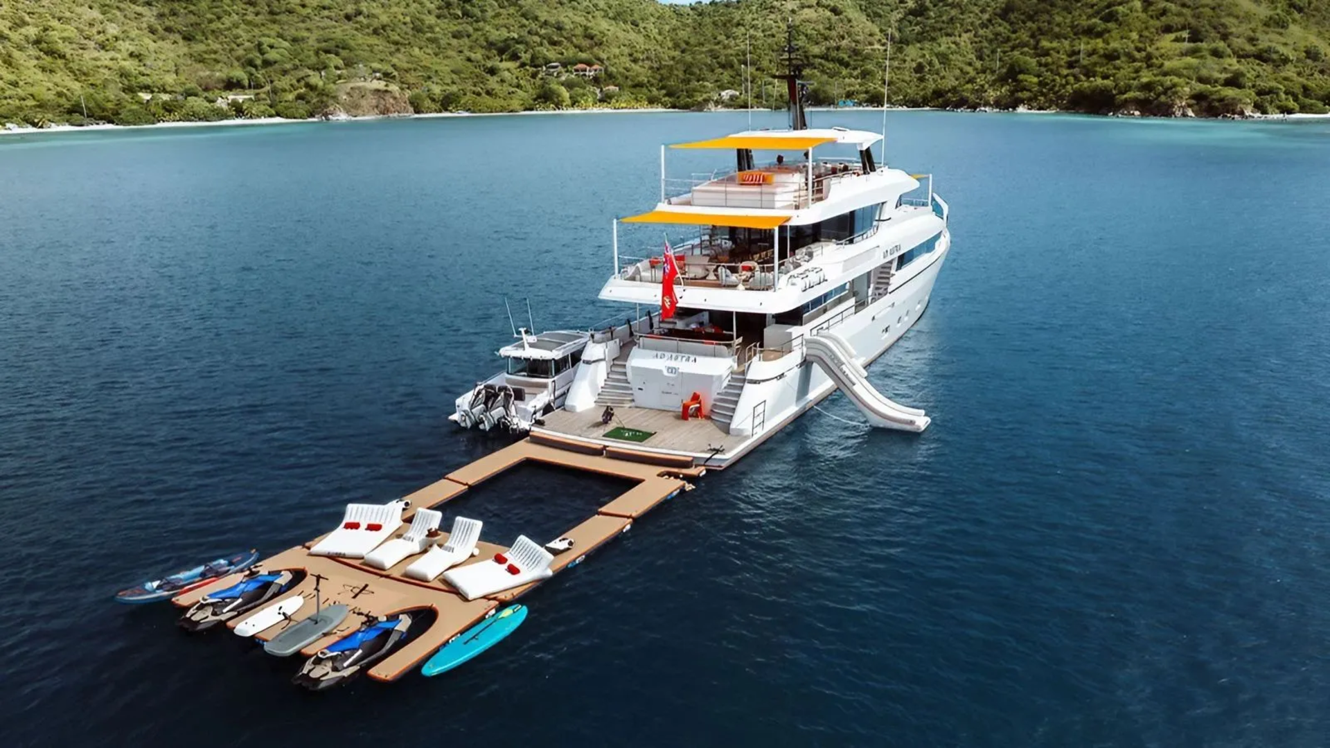 A large white yacht with a slide is docked at a floating platform with lounge chairs and water toys, set against a backdrop of lush green hills and calm blue water.