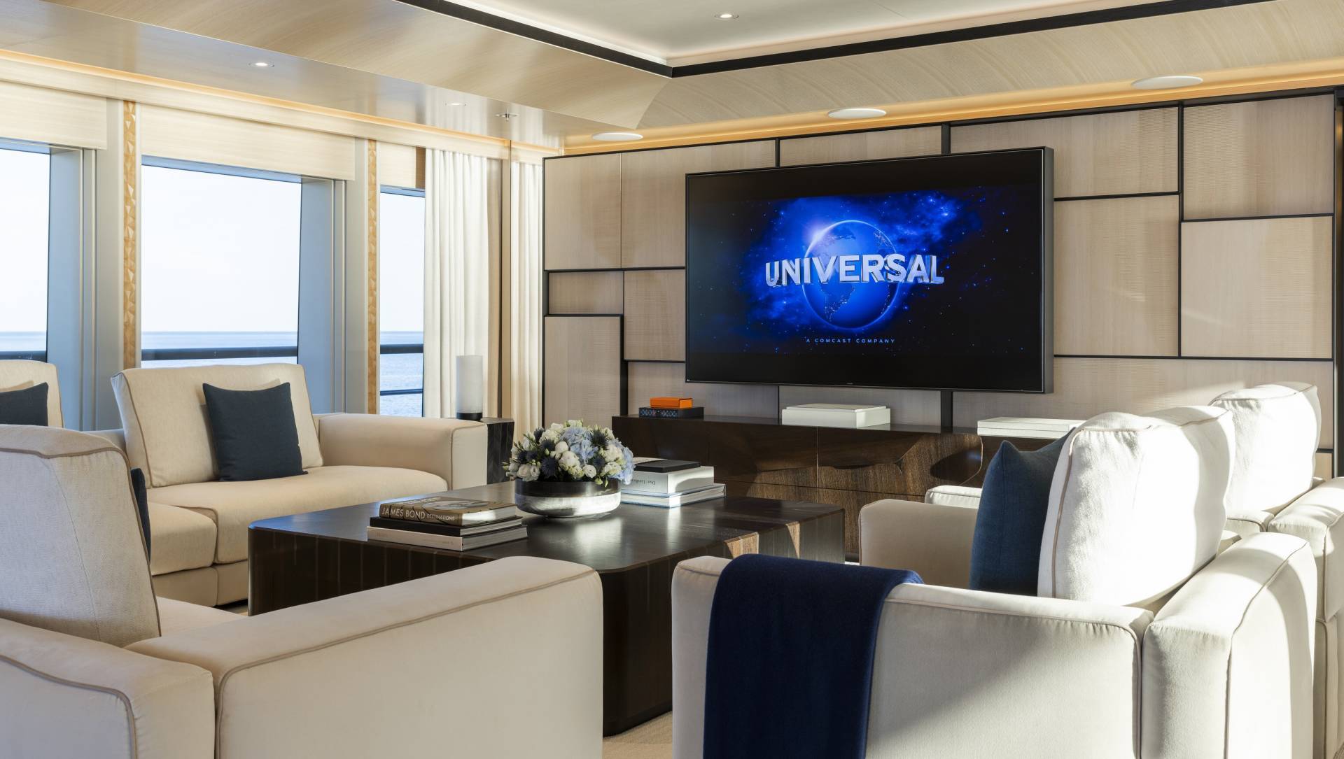 A plush living area with comfortable sofas and a large television displaying the Universal logo is set against a backdrop of ocean views.