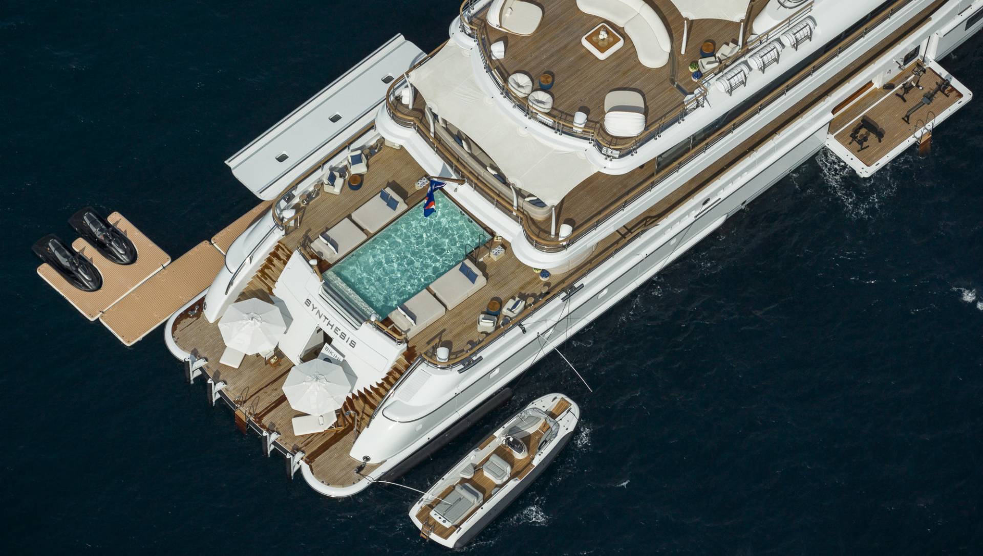 A luxurious yacht with a pool and lounge area is anchored in the deep blue sea, with a smaller tender boat nearby.