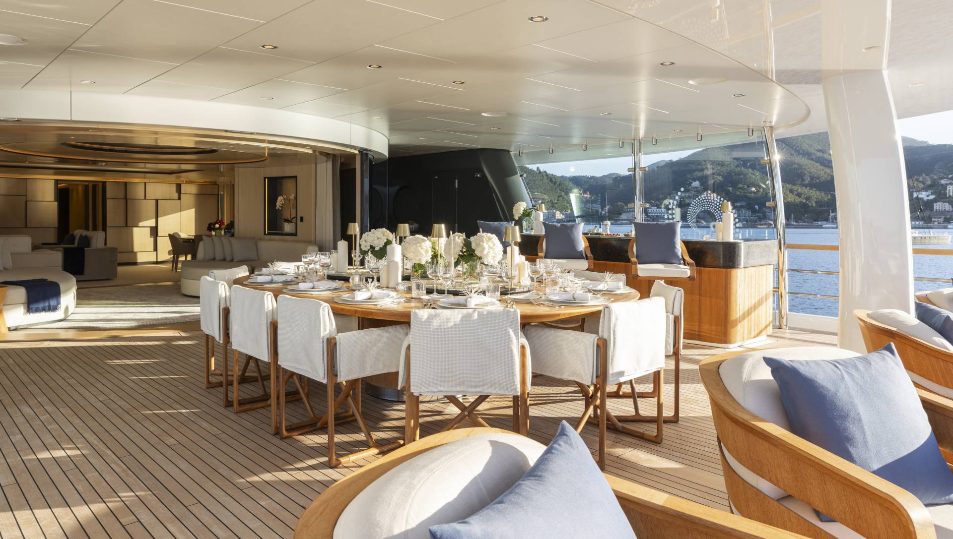 A luxurious yacht deck features a beautifully set dining table in the foreground, with comfortable seating and a scenic view of a sun-drenched coastline in the background.