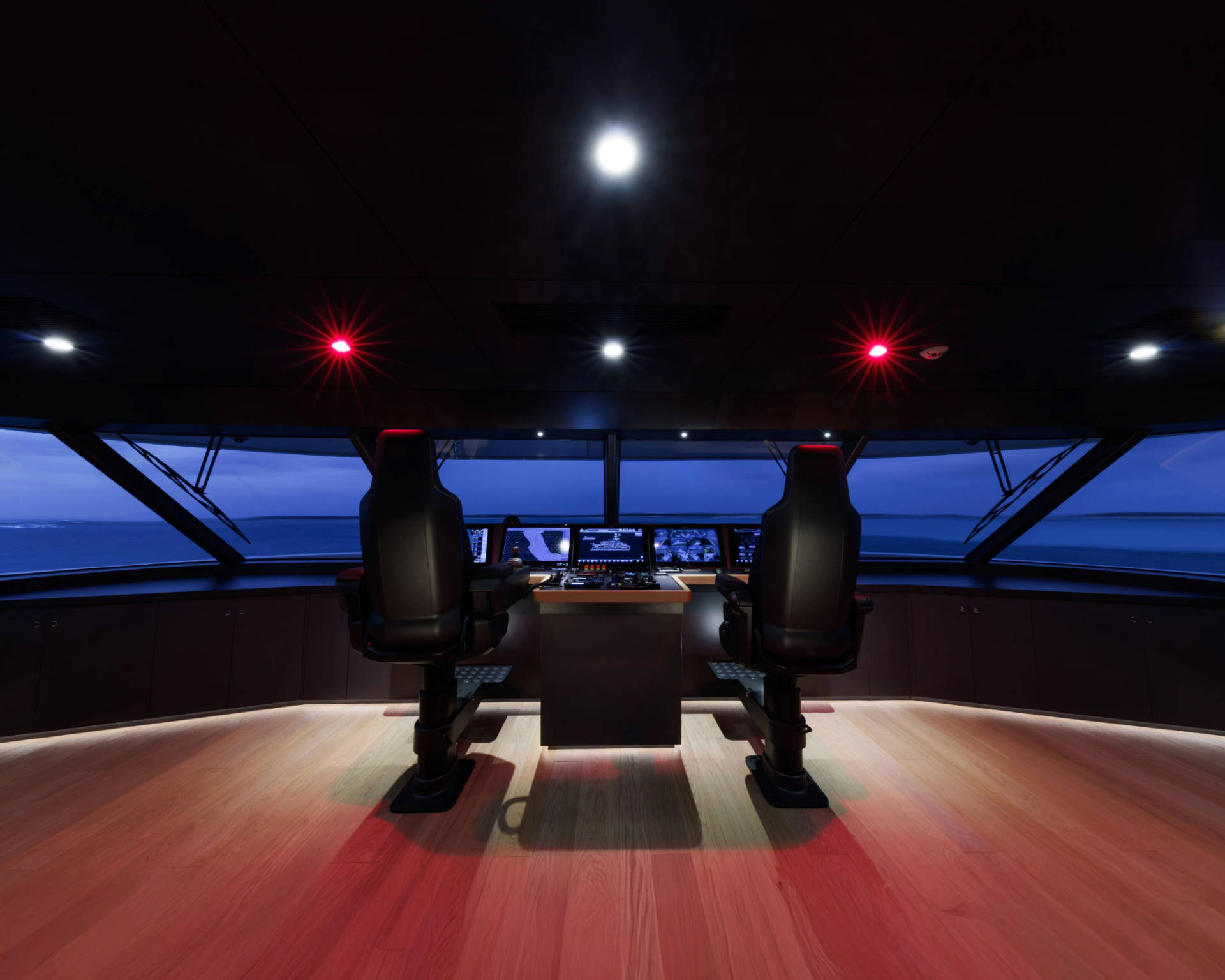 Two captain's chairs face a console of screens in a dimly lit yacht bridge, with the calm, blue ocean visible through the large windows.