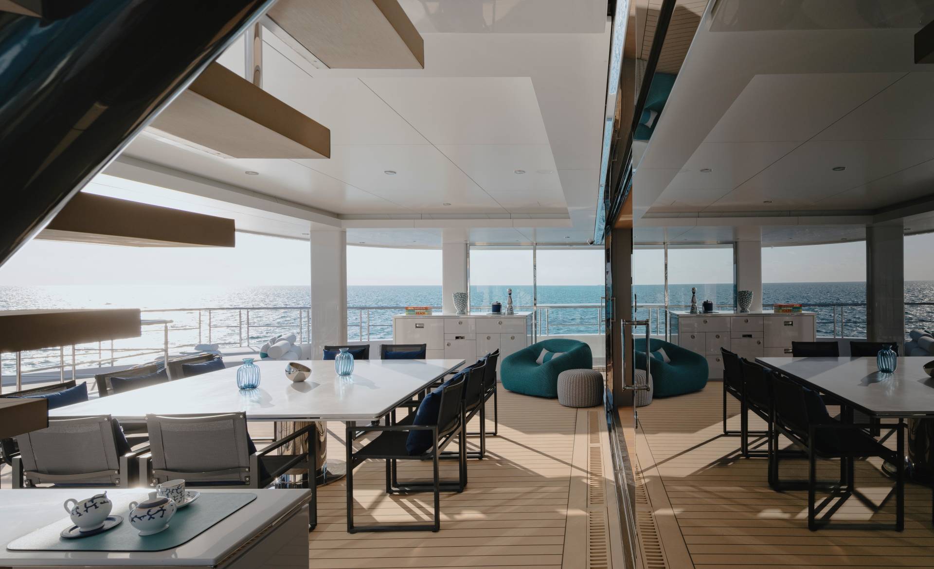 Modern outdoor dining areas with white tables and chairs are set against a backdrop of the calm ocean and a bright sky, reflected in a mirrored wall.