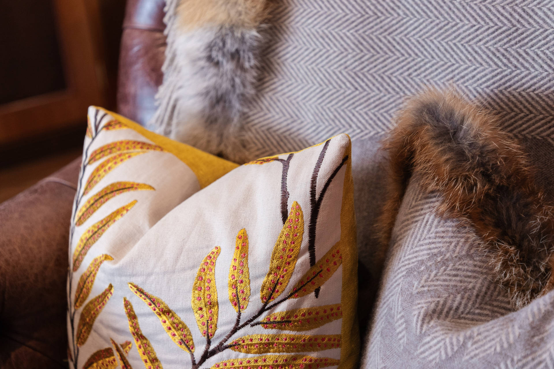 A cream pillow with a gold leaf pattern rests in the foreground against a background of a herringbone blanket and brown leather.