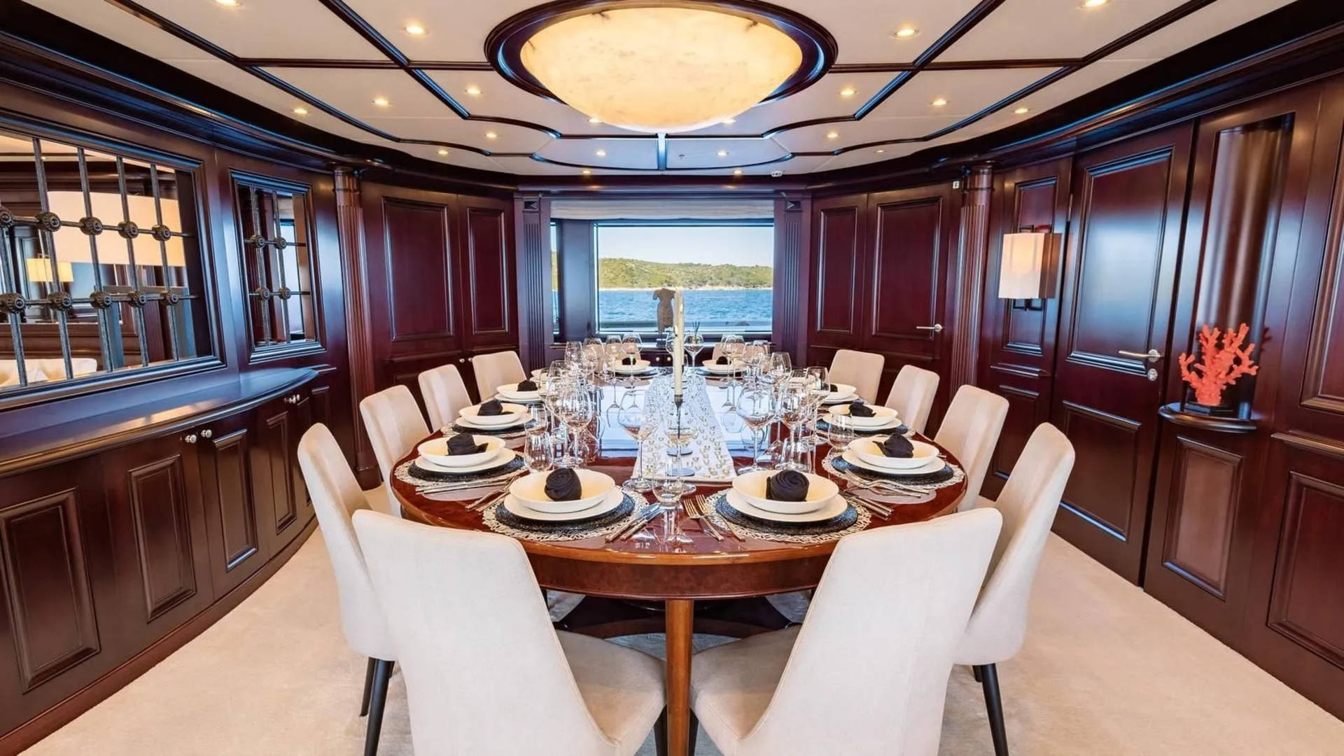 A formal dining table set for a feast is centered in a richly paneled room, with a large window in the background revealing a serene ocean view.