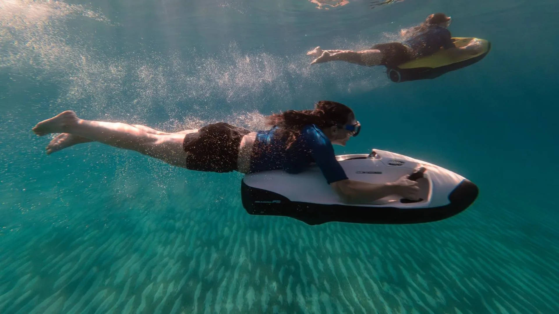 A woman glides through clear blue water on a sea scooter in the foreground, while another person on a similar device swims in the sandy background.