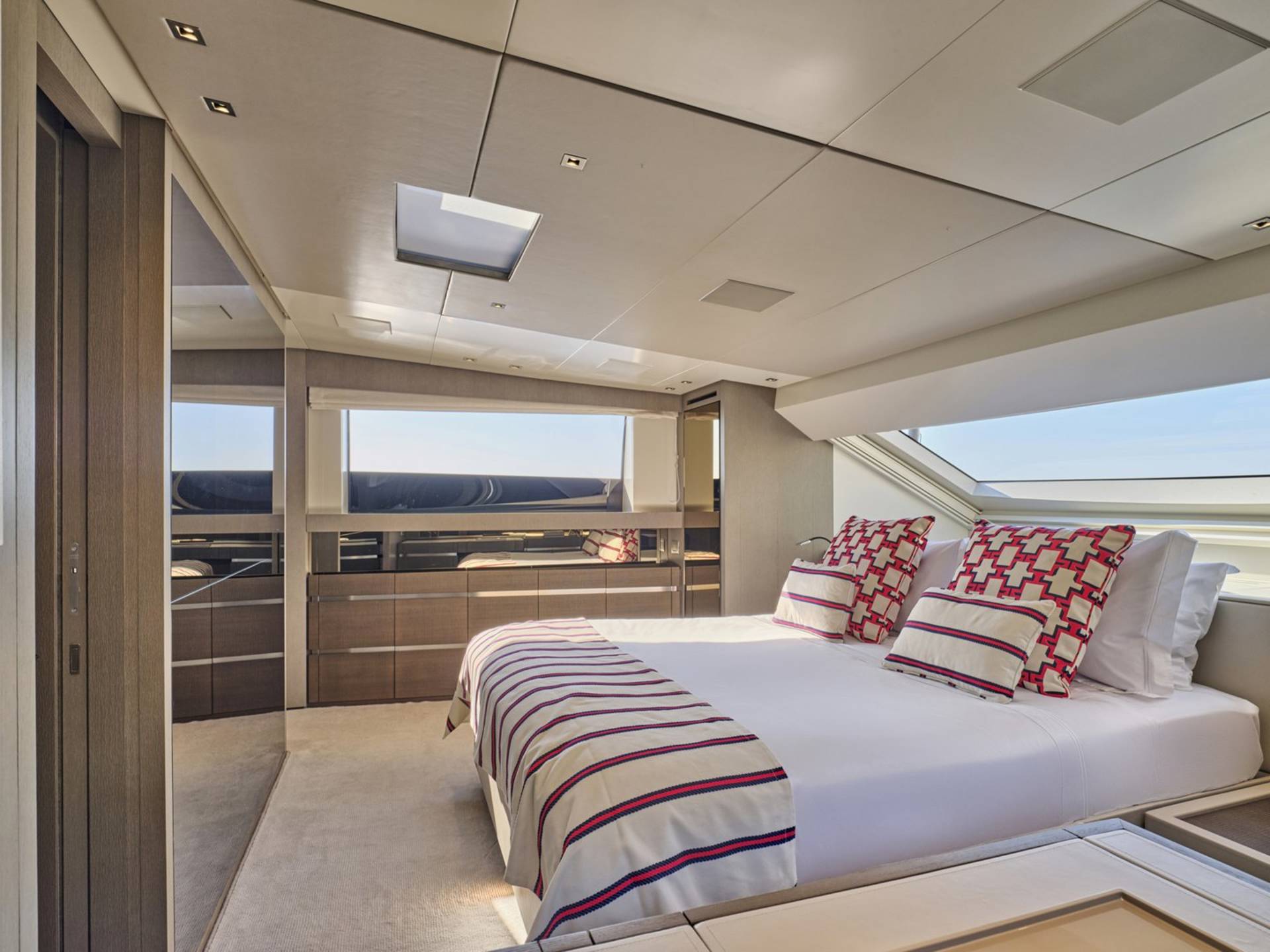 A luxurious yacht bedroom with a neatly made bed in the foreground and large windows offering a view of the sea in the background.