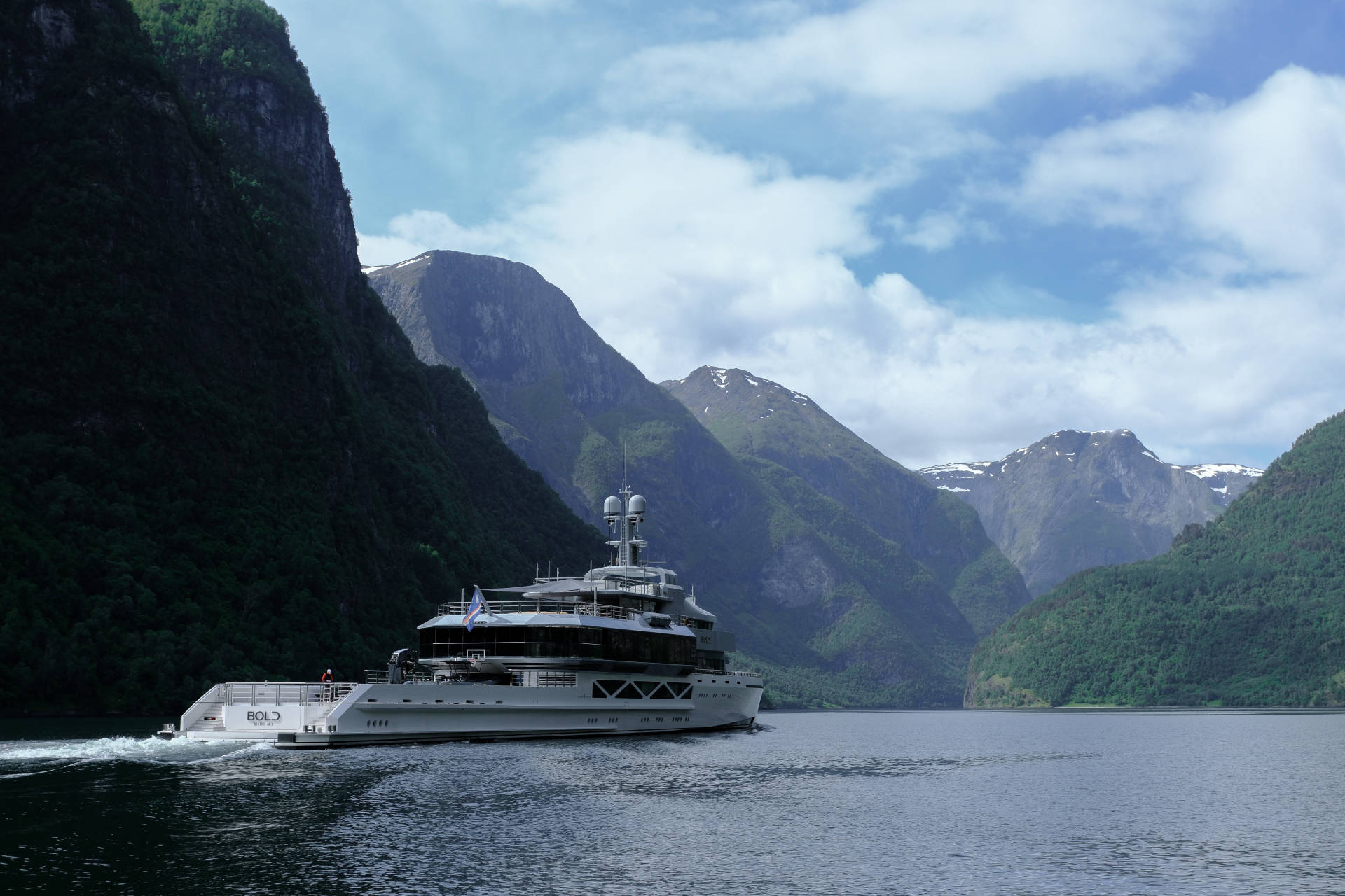 A large white yacht cruises through a calm body of water, dwarfed by the majestic, green-covered mountains that rise steeply on either side.