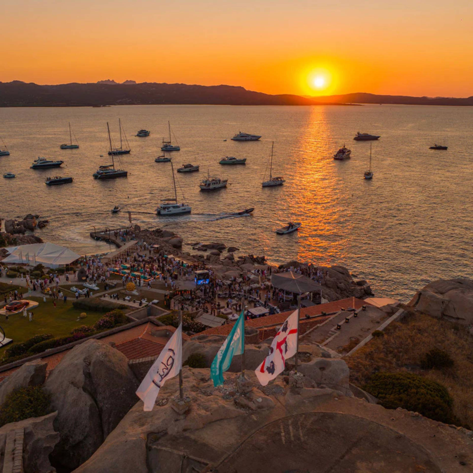 Flags wave in the foreground as a sunset illuminates a bay filled with boats and a lively gathering on the shore.