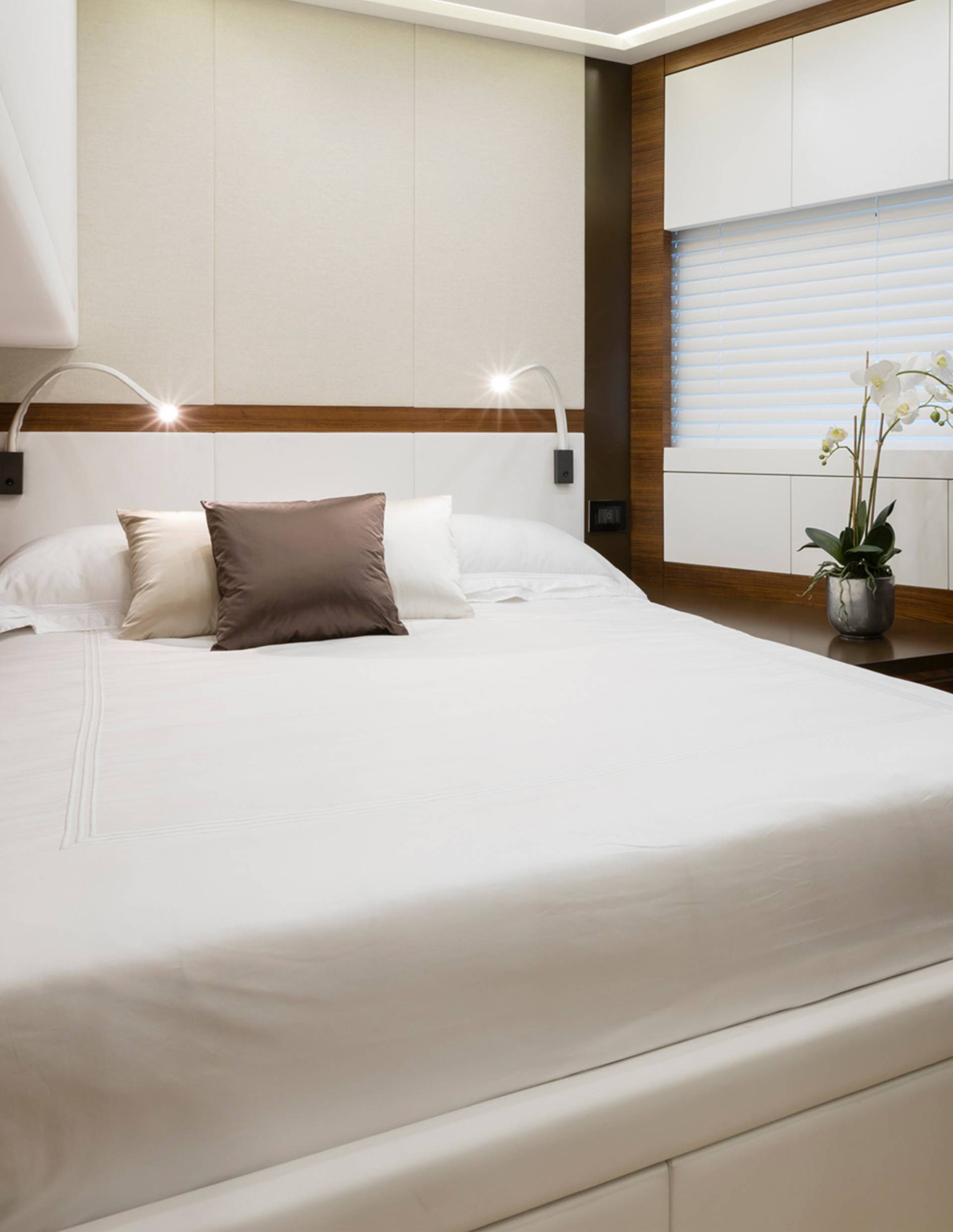 A neatly made bed with two pillows in the foreground is set against a wall with built-in cabinets and a window with blinds in the background.