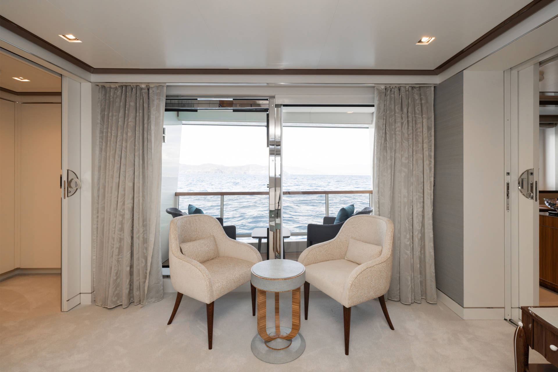 Two cream-colored armchairs flank a small round table in the foreground, with a balcony overlooking the ocean visible through large glass doors in the background.