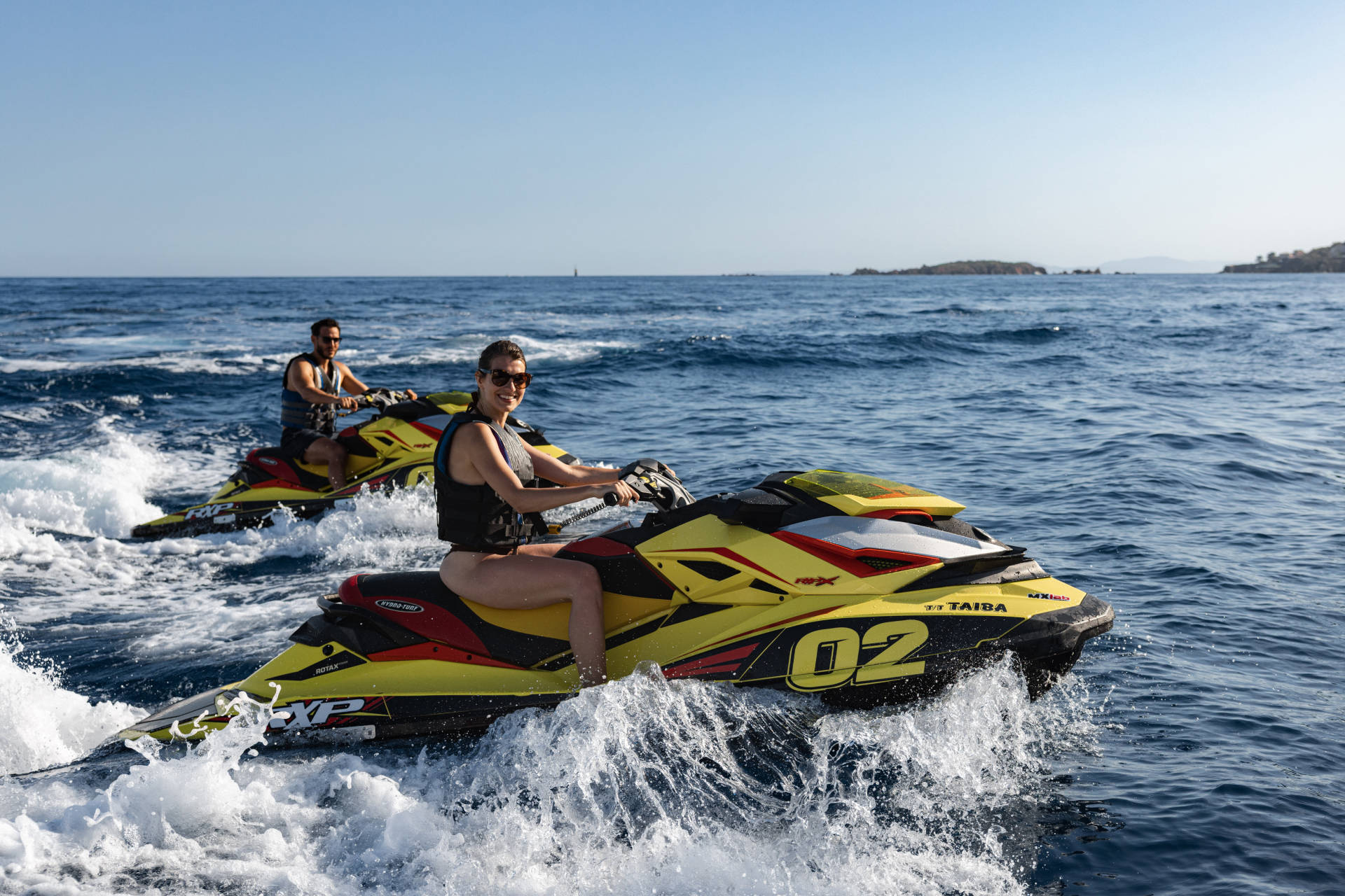 Two people ride yellow and red jet skis through choppy blue water, with a distant island under a clear sky.