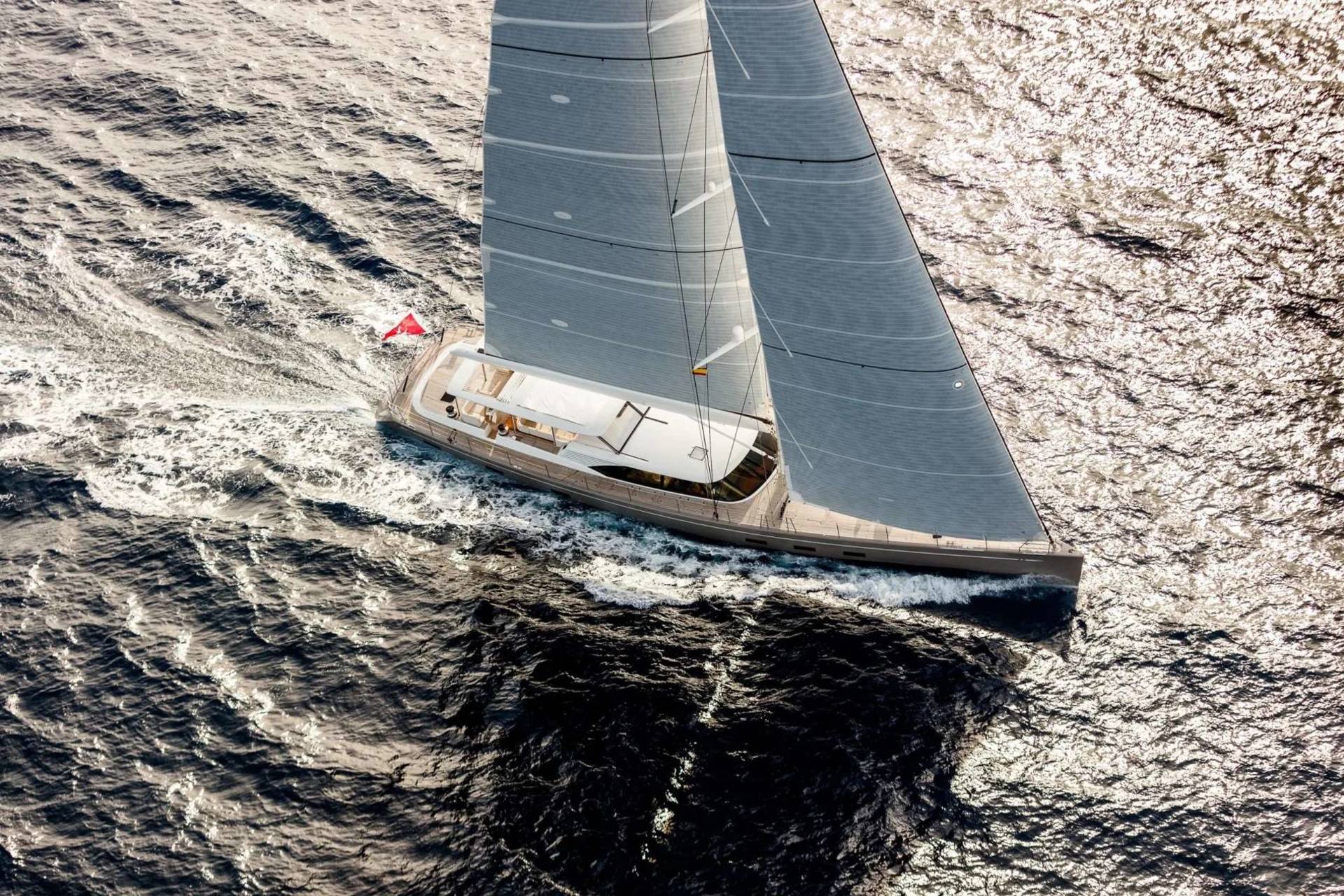 A sleek sailboat with grey sails cuts through choppy, dark water, its shadow stretching out behind it against a sunlit sea.
