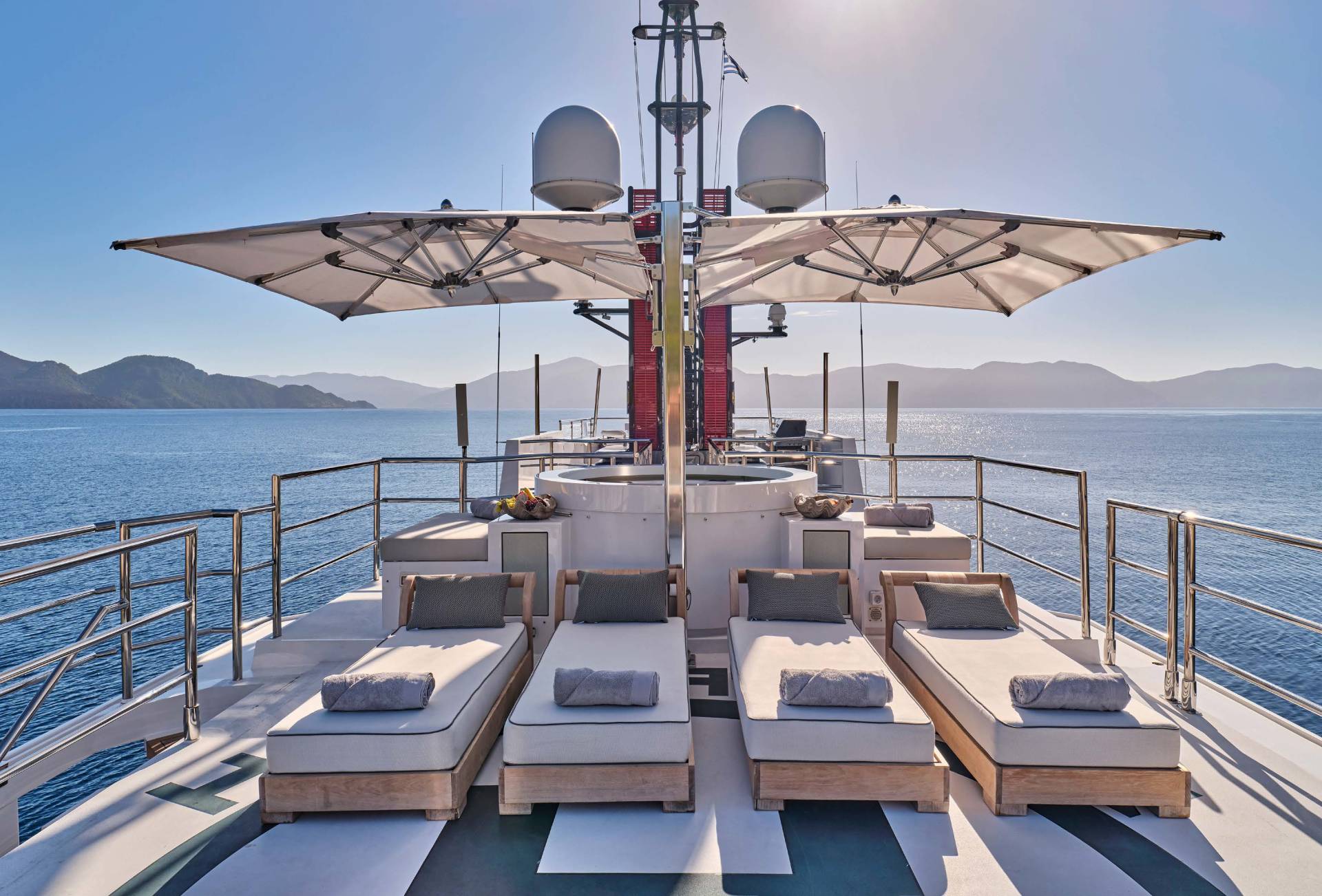 Four sun loungers with grey cushions are arranged on the deck of a yacht, with a clear blue sky and distant, hazy mountains in the background.