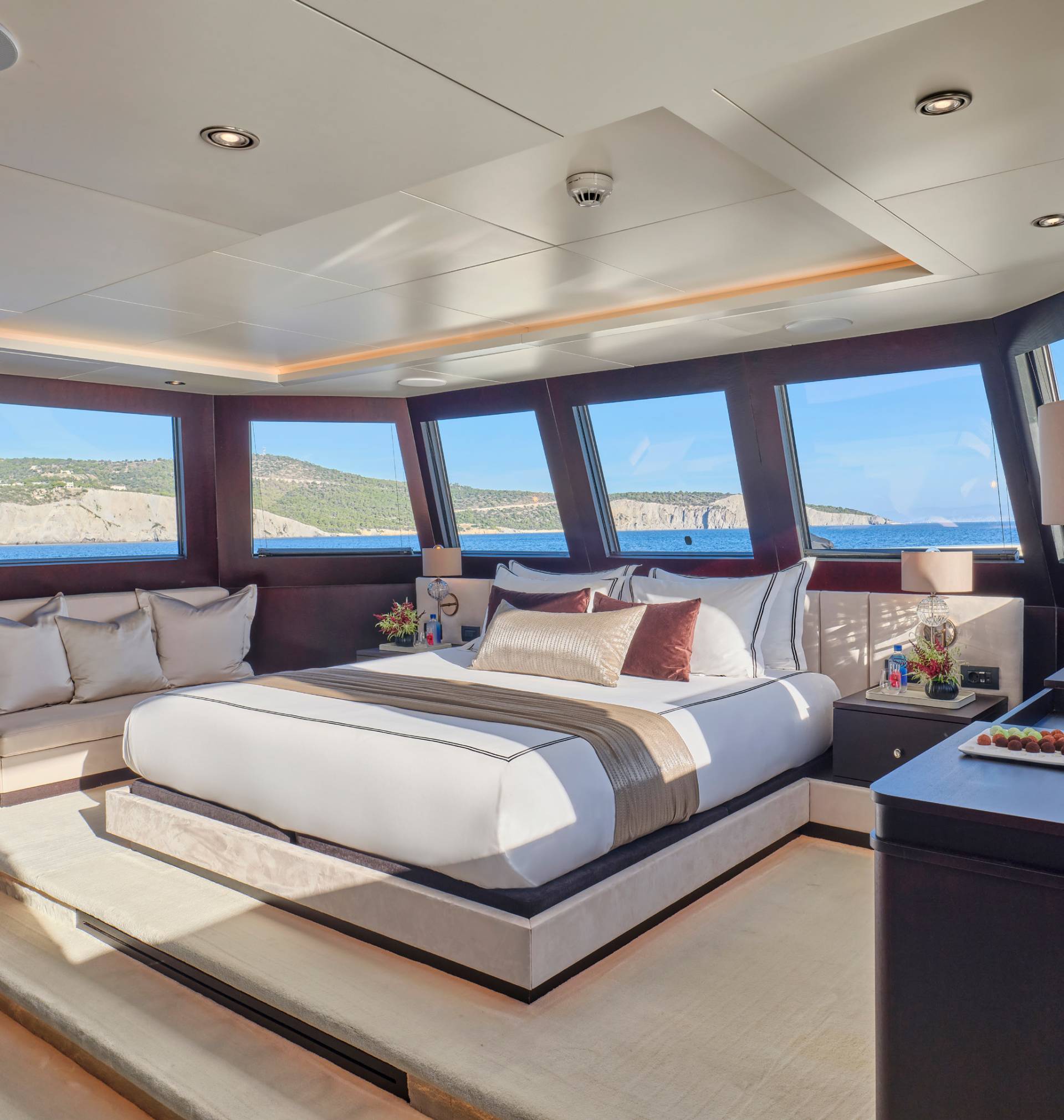 A luxurious bedroom with a large bed and sofa in the foreground is set against a backdrop of a scenic coastal view through large windows.