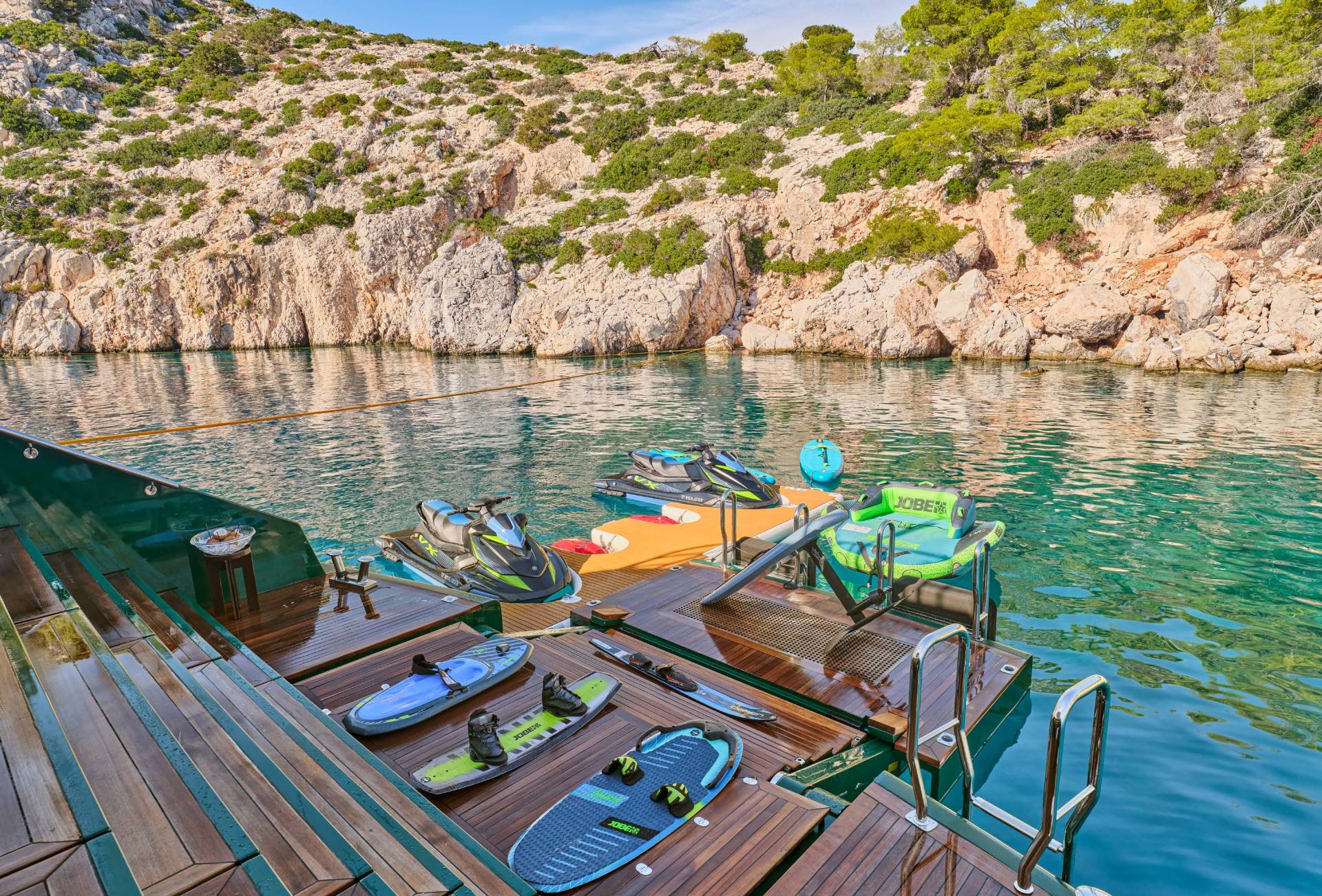 Jet skis and wakeboards rest on a wooden dock in the foreground, with a rocky, tree-covered coastline and calm turquoise water in the background.