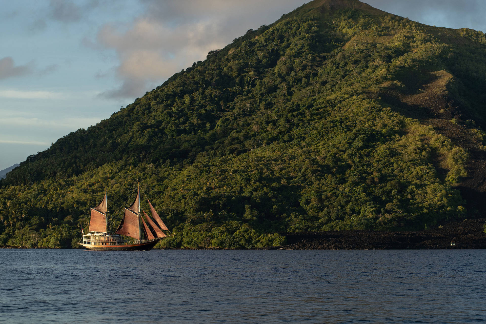 A wooden sailing ship with reddish-brown sails glides across dark blue water in the foreground, with a lush green, tree-covered mountain rising steeply in the background.