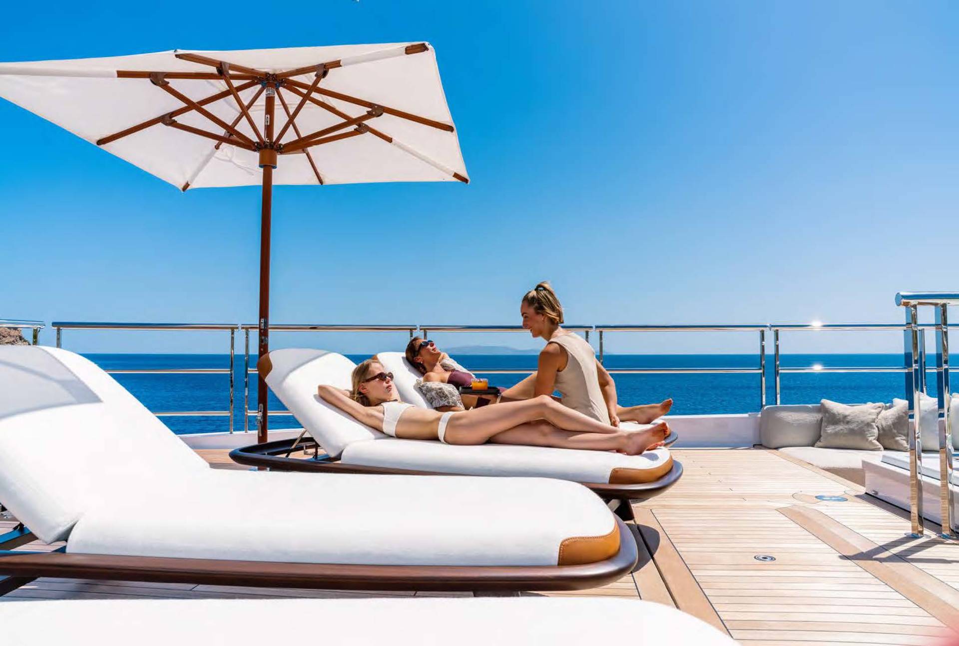 Three people relax on lounge chairs on a yacht's deck under a large umbrella, with the blue ocean stretching to the horizon under a clear sky.
