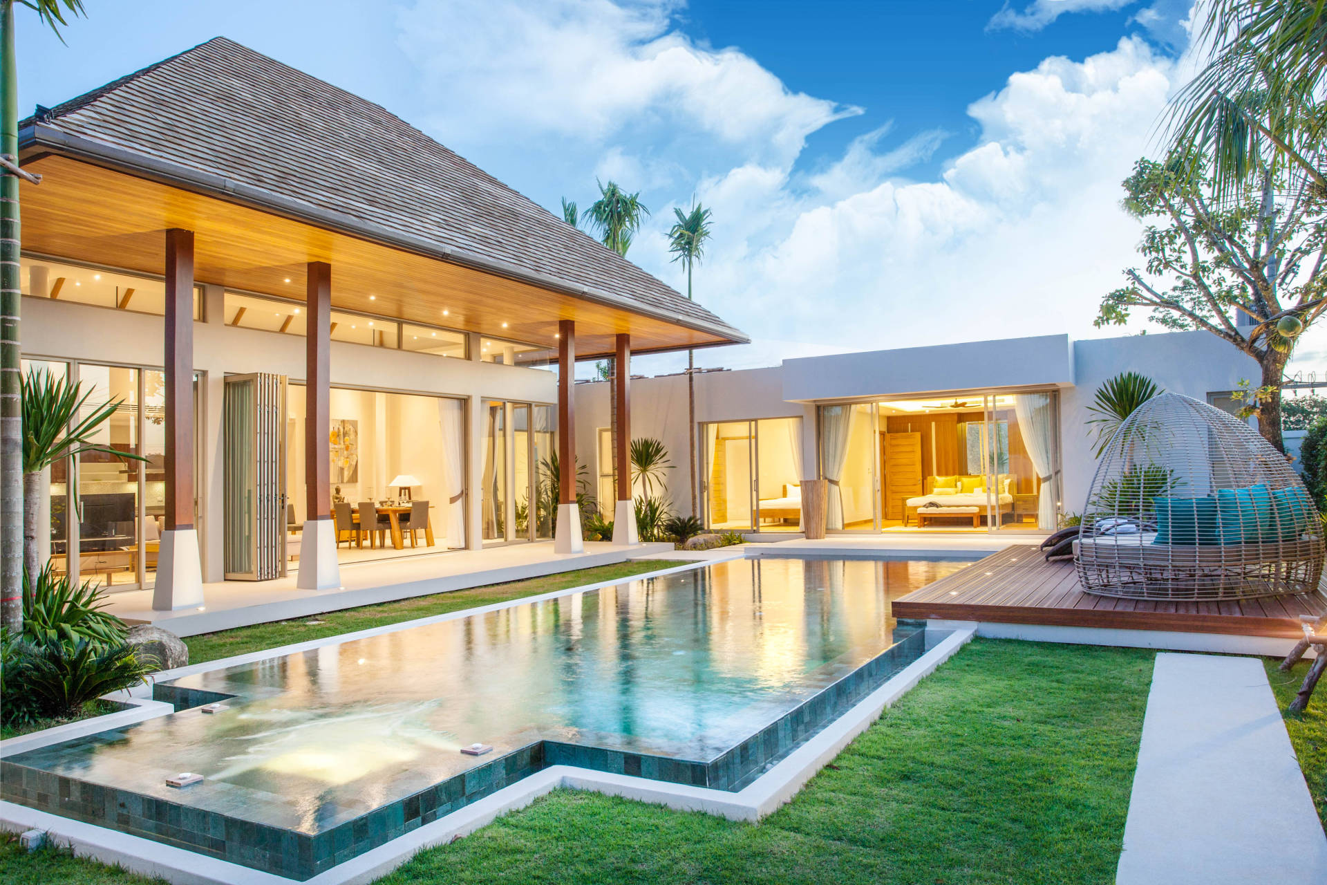A modern villa with a swimming pool in the foreground is set against a backdrop of a bright blue sky with fluffy clouds.