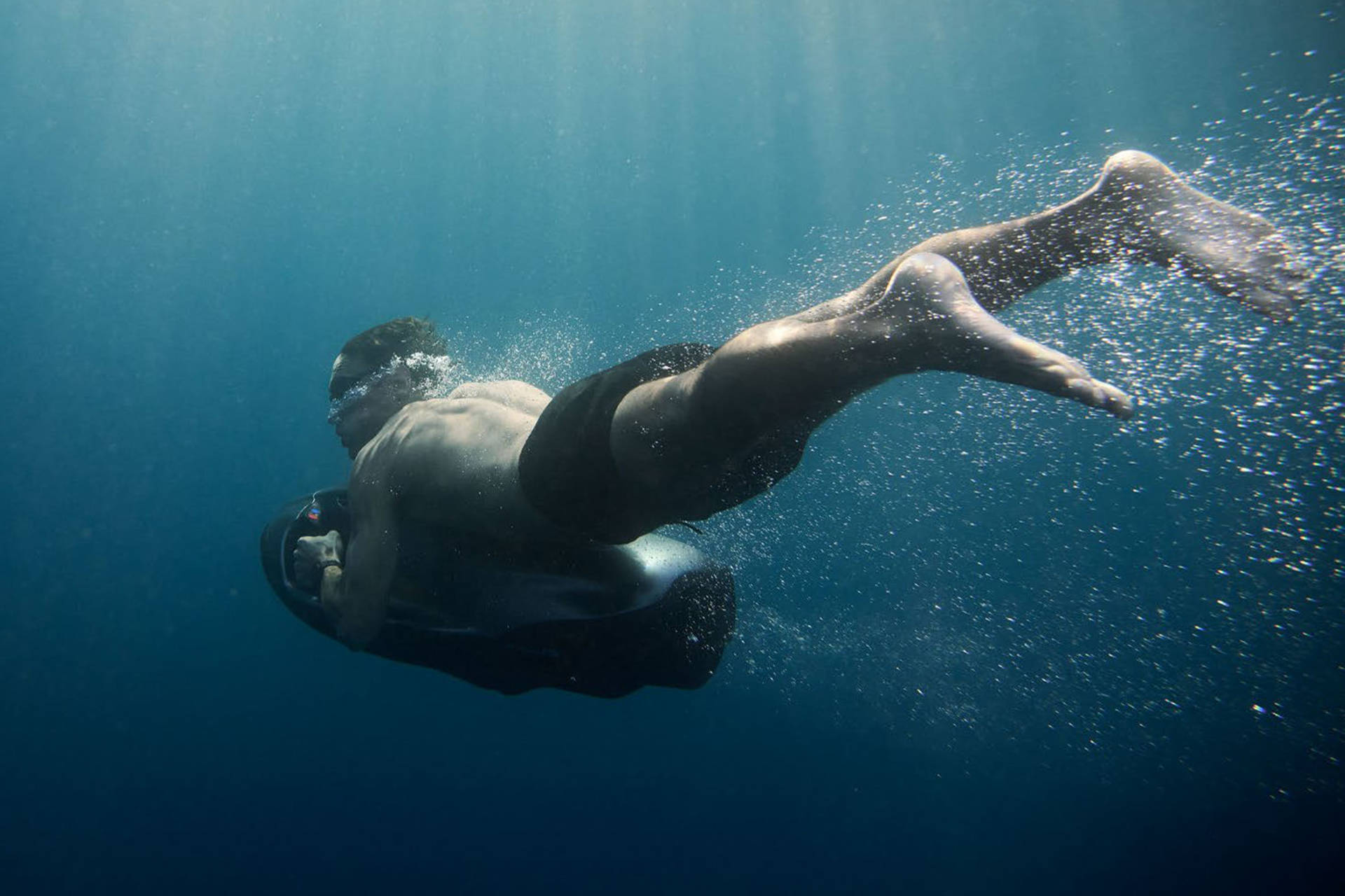 A man with goggles and black shorts propels himself through clear blue water with a sea scooter, leaving a trail of bubbles against a deep blue background.