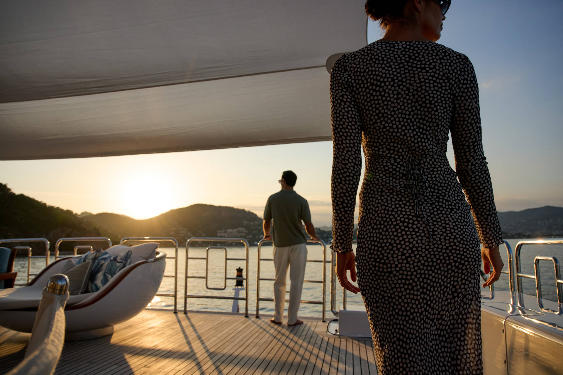 A woman in a patterned dress stands on a yacht's deck in the foreground, while a man gazes at the sunset over a hilly coastline in the background.