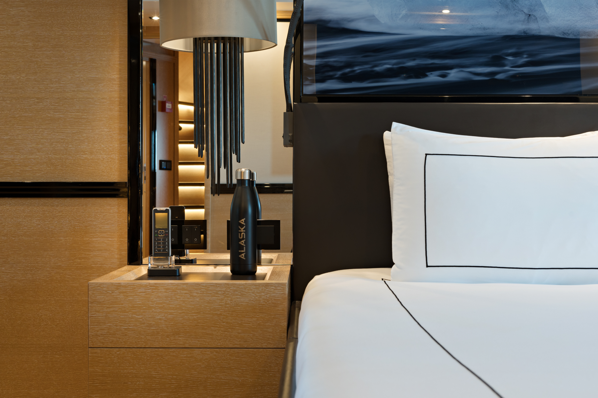 A modern bedroom features a bedside table with a phone and water bottle in the foreground, and a large television displaying a scenic landscape in the background.