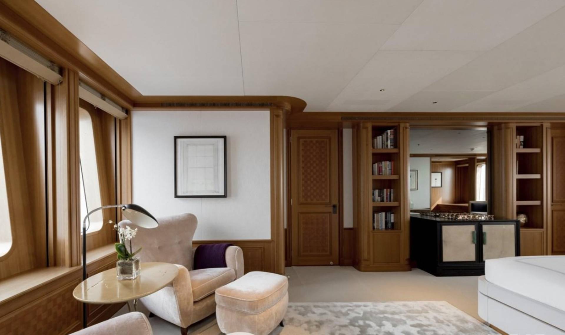 A plush armchair and ottoman sit in the foreground of a well-lit room with large, curved windows, while a built-in bookshelf and bar area are visible in the background.