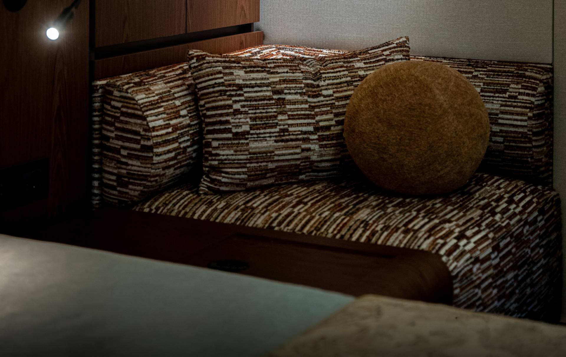 A cozy seating area with patterned cushions and a round textured pillow is illuminated by a spotlight against a dark wooden background.