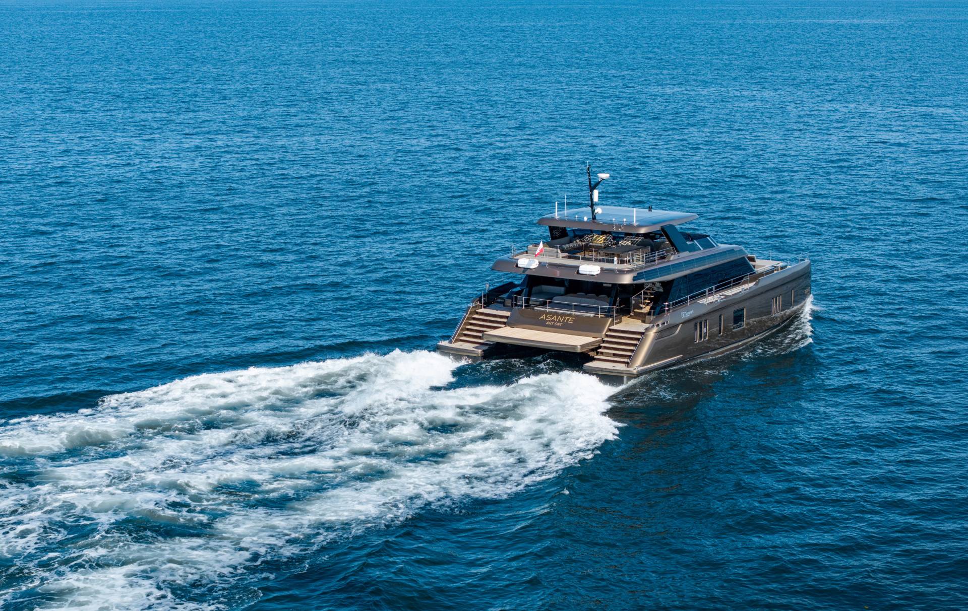 A sleek, modern catamaran yacht cruises through calm blue waters, leaving a white wake behind it against a clear sky.