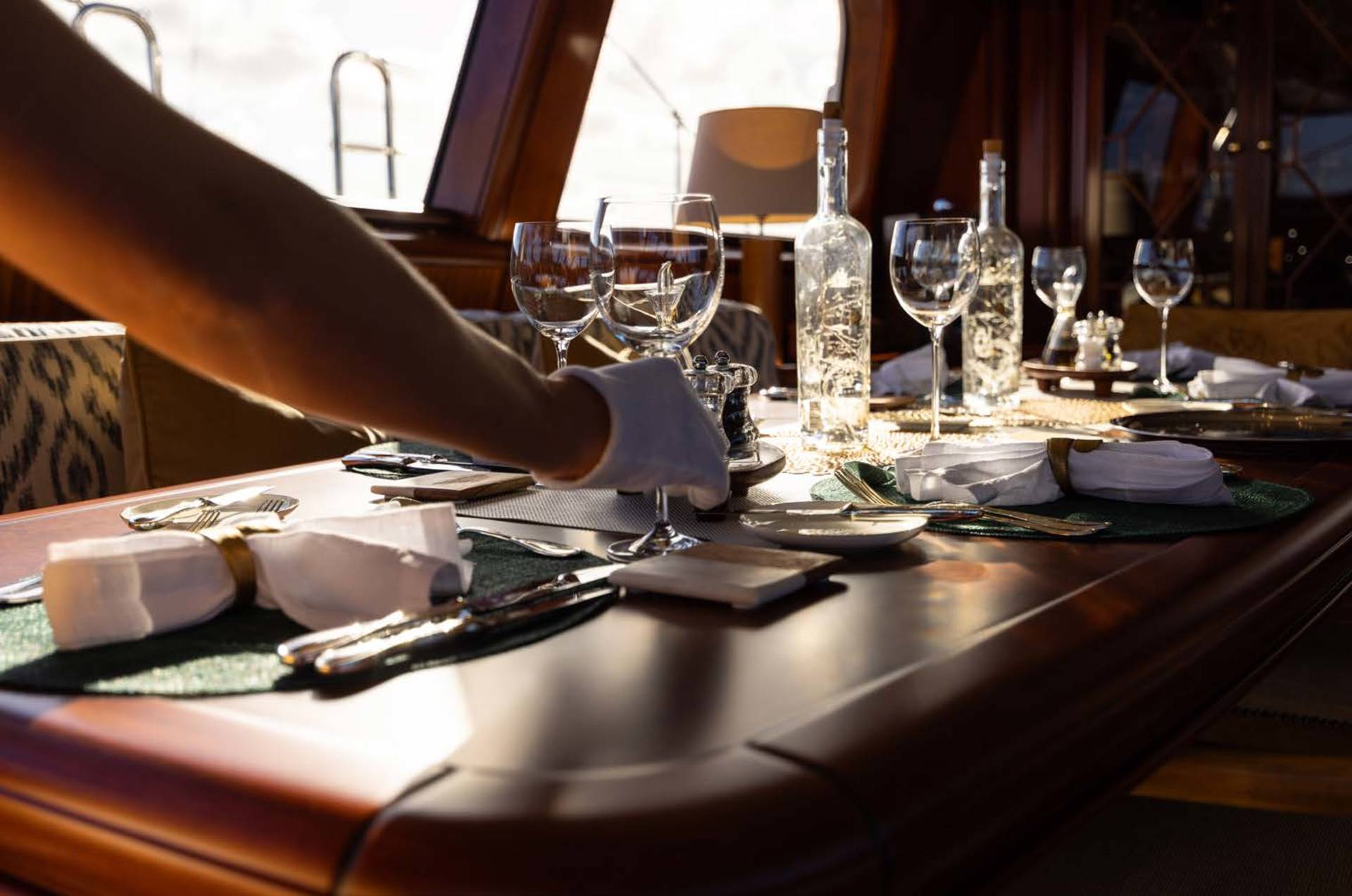 A gloved hand seasons a beautifully set dining table on a yacht, with bottles of water and wine glasses arranged on the polished wood surface, while sunlight streams in from the windows in the background.